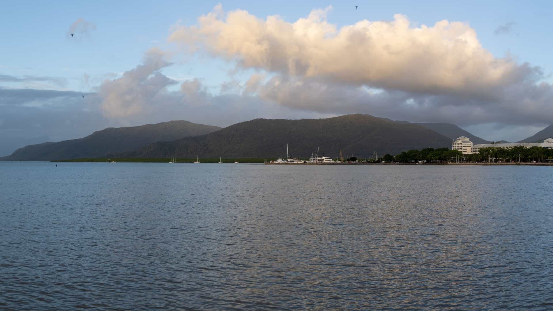 A serene view of the calm waters along the Cairns Esplanade in Queensland, Australia. The waterfront features a public swimming lagoon, with palm trees and a boardwalk visible. The city skyline is in the background.