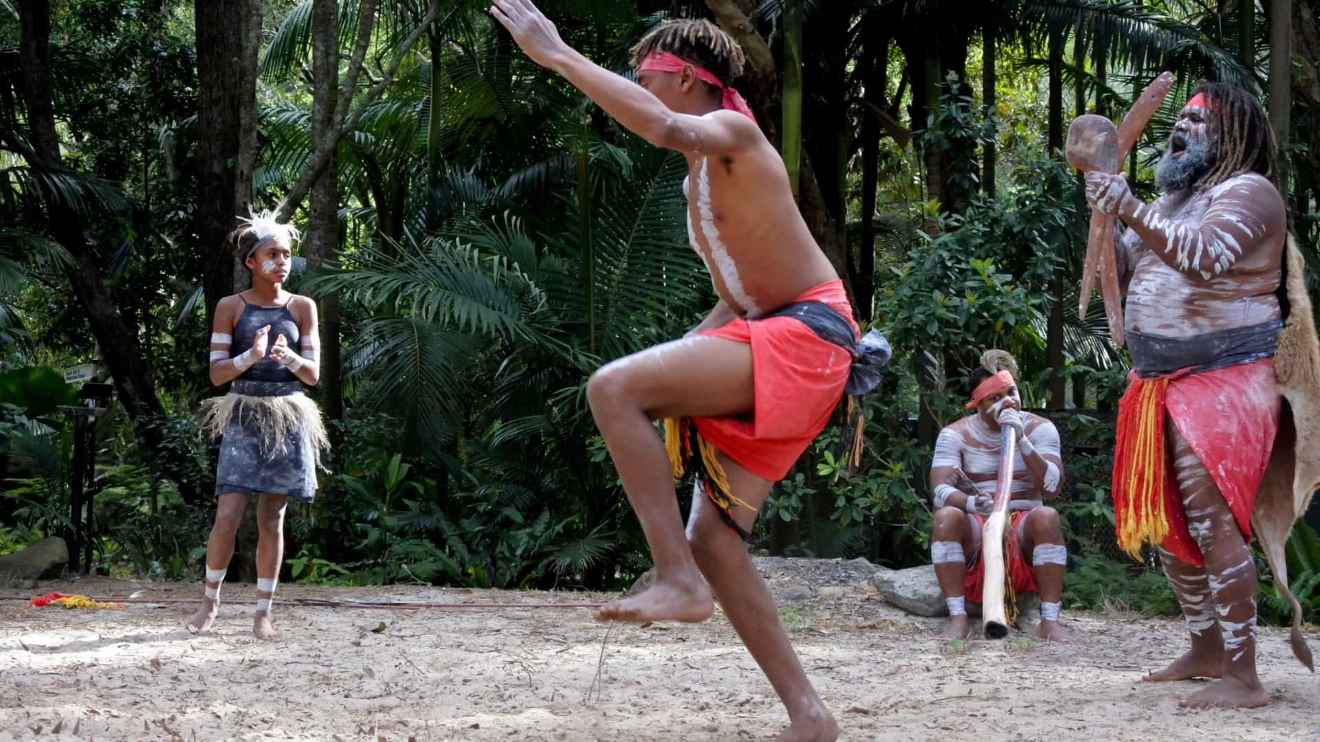 Aboriginal Culture at Tjapukai Aboriginal Cultural Park, featuring a dancer with traditional body paint and attire performing a ceremonial dance on a stage in a natural, open air or rainforest like setting.