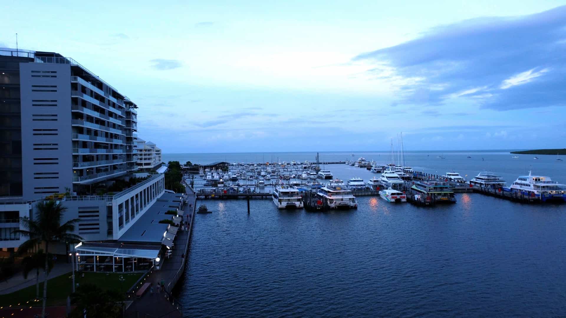 A bustling photo of the Cairns marina in Queensland, Australia, showcasing various boats docked at the port with the city's modern high rise buildings and waterfront hotels in the background.