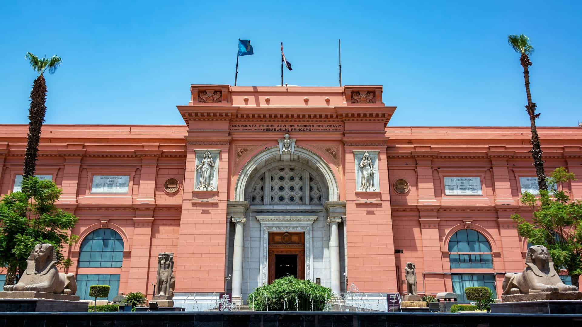 An exterior view of the Egyptian Museum in Cairo, Egypt, showcasing its neo classical Beaux Arts architecture with a central archway flanked by columns and statues. The surrounding area includes palm trees and a street with cars.