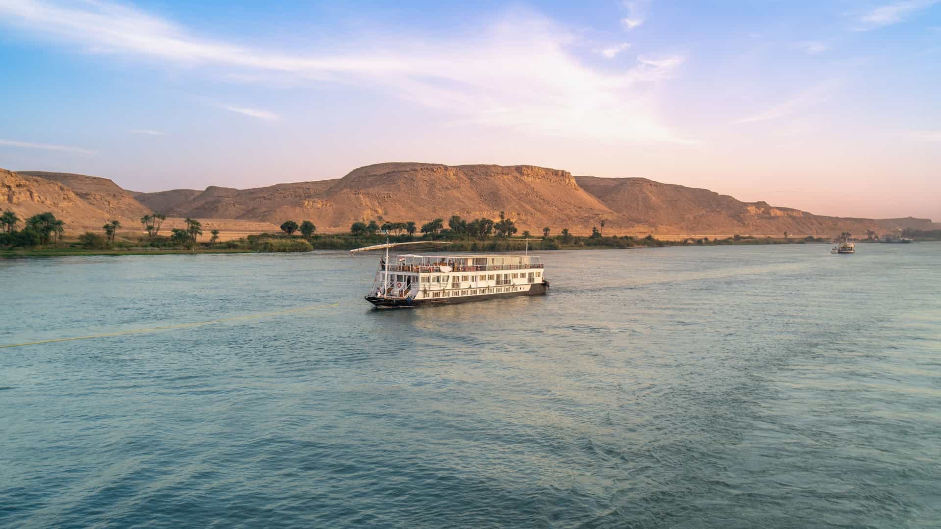A serene scene on the Nile River near Aswan, Egypt, with two cruise boats and a smaller sailboat on the water during a tranquil dusk. The sky is a soft mix of colors, reflecting on the calm river.