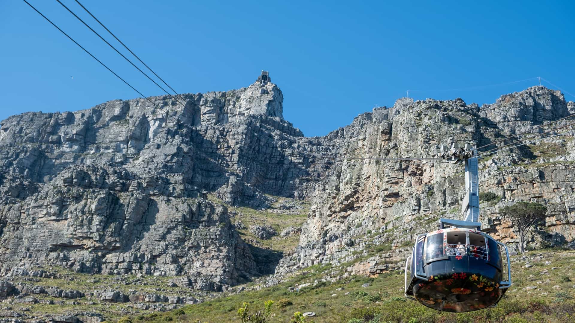 A modern Table Mountain Aerial Cableway car, with a large viewing window, ascends the rocky face of Table Mountain in Cape Town, South Africa. The car is set against a backdrop of a clear blue sky, with a sliver of the distant city visible below.