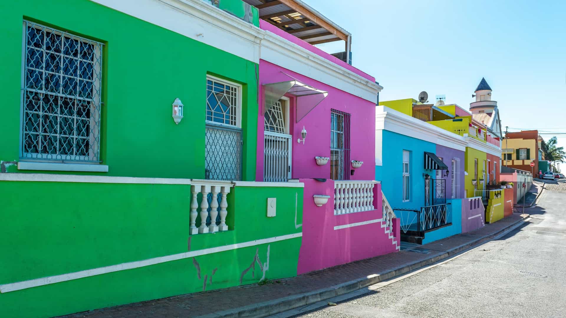 A vibrant street view in the Bo Kaap neighborhood of Cape Town, South Africa, featuring a row of brightly painted houses in various colors, including pink, yellow, and blue. The houses are located on a sloping cobblestone street, conveying a sense of historical and cultural significance.