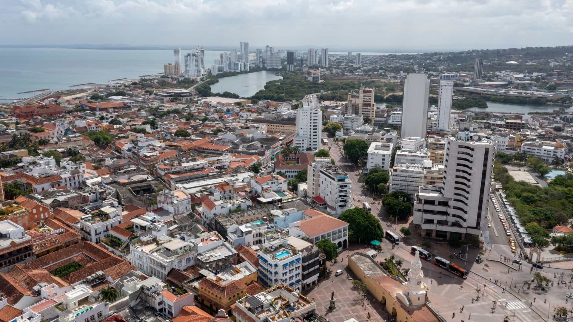 A view of the historic Plaza de los Coches in the walled city of Cartagena, Colombia. The image shows the plaza with its iconic archways and the prominent clock tower (Puerta del Reloj) in the background.