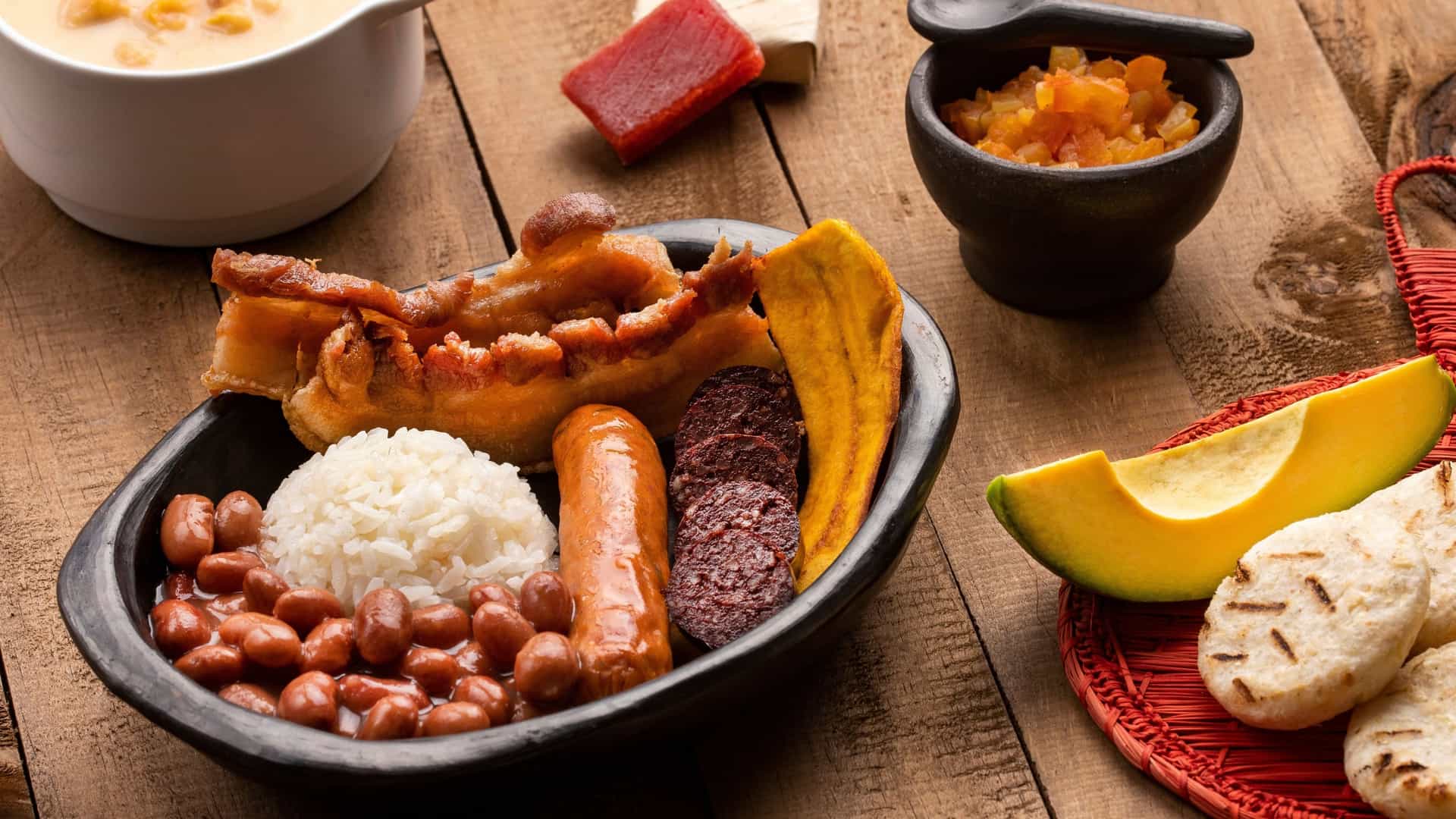 This vibrant photo captures a massive plate of Bandeja Paisa, the traditional Colombian platter from the Antioquia region. The hearty meal is an assortment of ingredients, including rice, red beans, ground meat, a fried egg, chorizo sausage, chicharrón (fried pork belly), fried plantain, and a slice of avocado, all artfully arranged on a large platter.