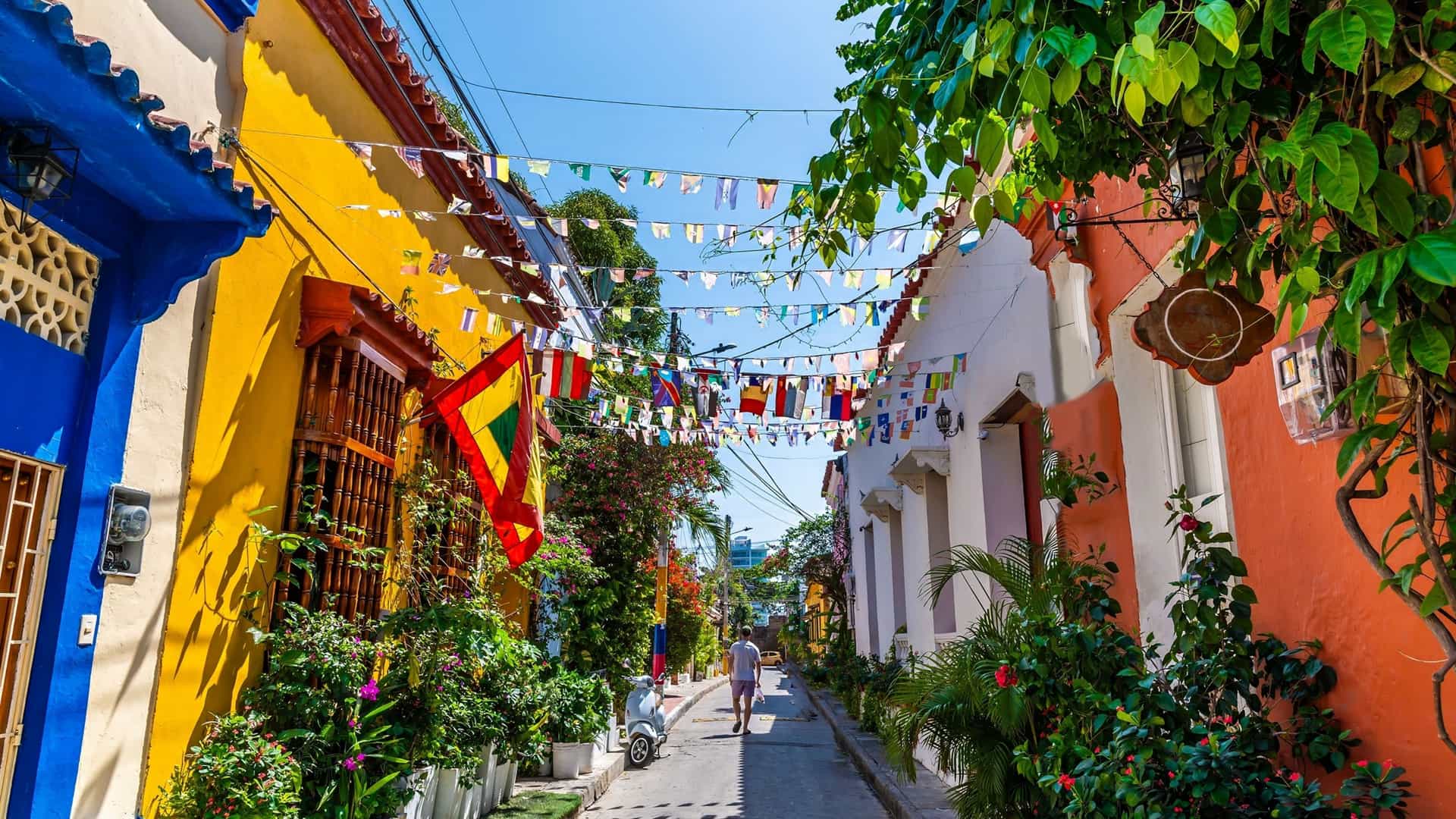 This stunning image shows a narrow, cobblestone street in the colonial city of Cartagena, Colombia, lined with brightly painted walls. The view is dominated by an ornate, flower adorned balcony overflowing with vibrant red and pink bougainvillea.