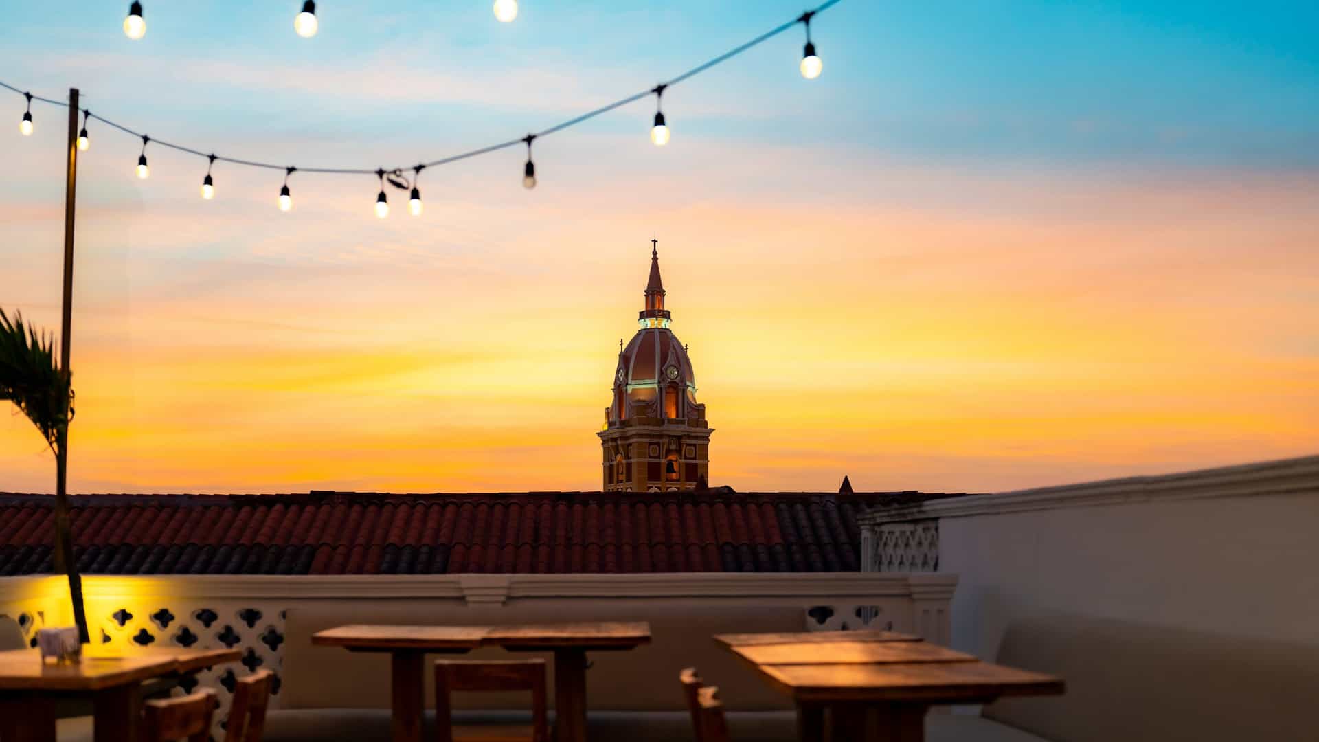 This stunning Sunset at Café del Mar view captures the illuminated dome of the San Pedro Claver Church, silhouetted against a breathtaking orange and turquoise sky. The image is taken from a charming rooftop terrace in Cartagena, featuring stylish tables and string lights, perfect for an evening dining experience.