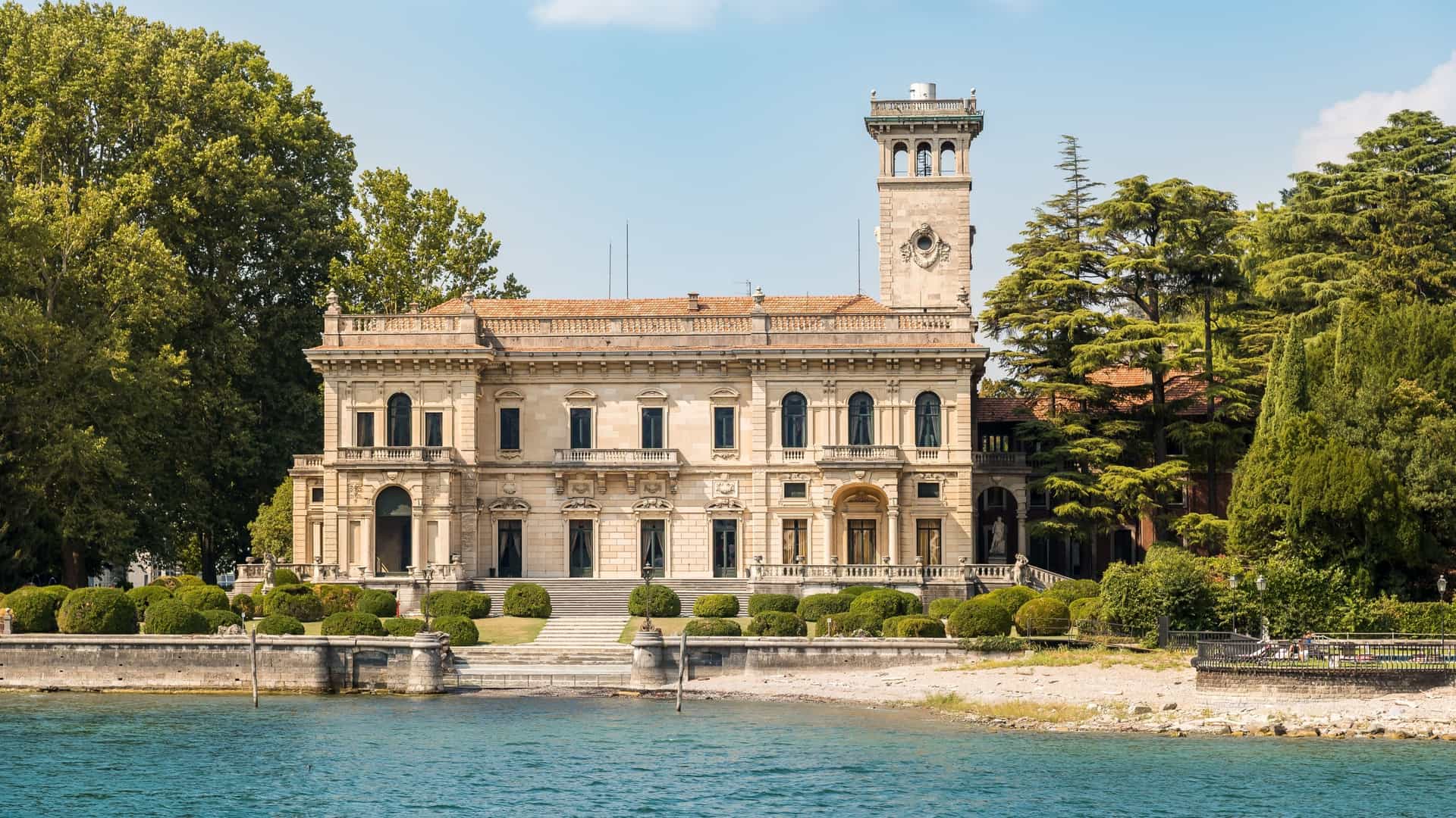 A wide angle view of Villa Erba on Lake Como, showcasing the magnificent 19th century Italian Mannerist style mansion with its imposing square structure, ornate facade, and central turret. The villa is surrounded by lush green gardens that slope down to the tranquil blue water of the lake.