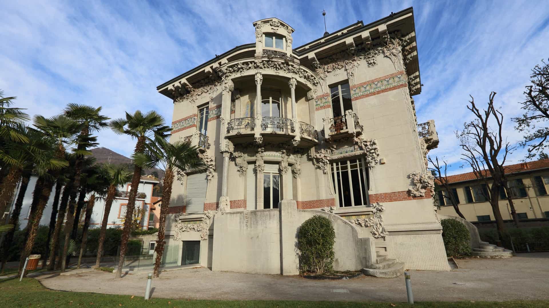 A close up view of Villa Bernasconi, a yellow Art Nouveau mansion on Lake Como, distinguished by its asymmetric structure, central turret, and curved balconies. The facade features elaborate sculptural reliefs depicting the silkworm life cycle (silkworms, mulberry leaves, and butterflies), a tribute to the owner's silk industry wealth.