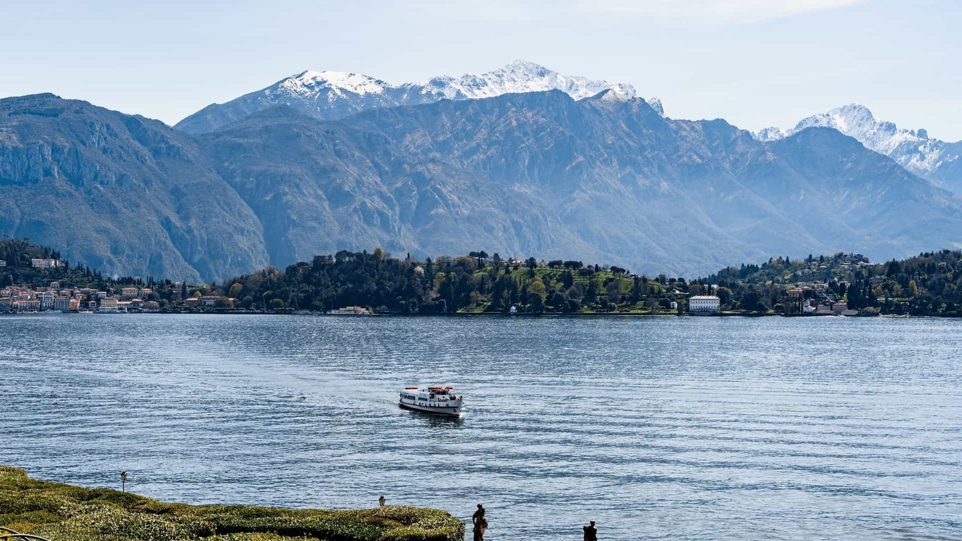 A ferry sails on the serene waters of Lake Como, Italy, with snow covered mountains in the background.