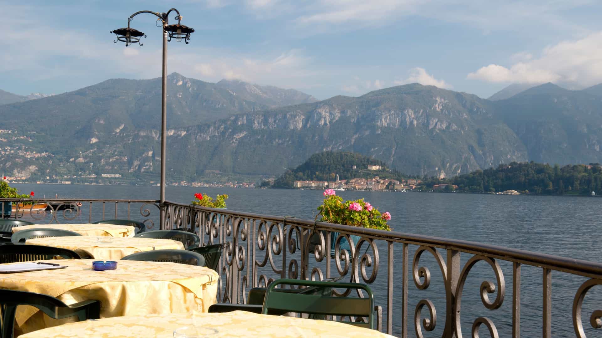 A scenic view of an upscale restaurant terrace on the shores of Lake Como, Italy, featuring white tablecloths, comfortable chairs, and a wisteria covered or pergola roof. The dining area overlooks the calm blue waters of the lake, with lush, green mountains rising steeply in the background.