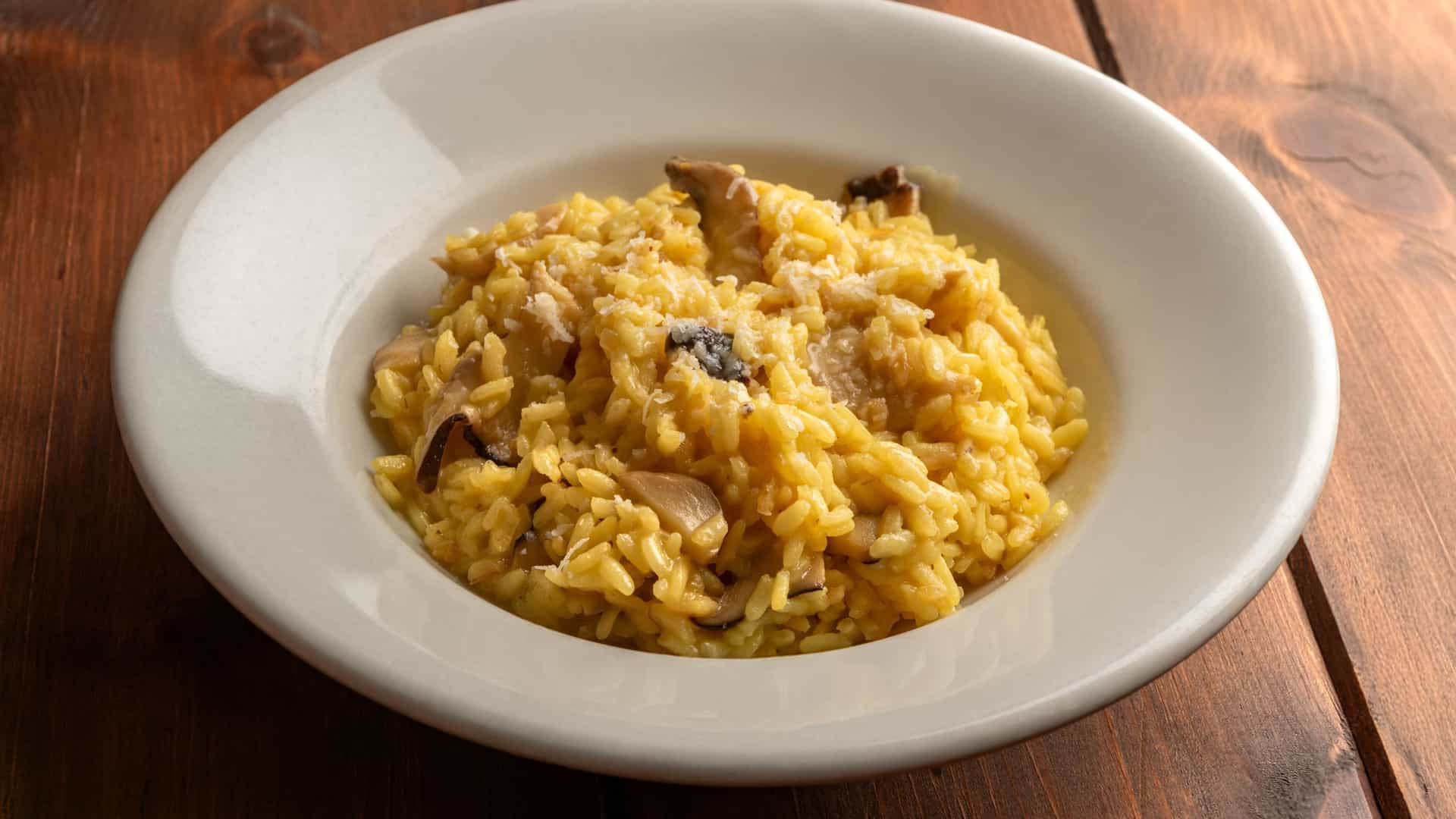A close up, top down view of a single serving of Italian mushroom risotto on a white plate. The creamy rice is visibly speckled with sliced mushrooms (likely porcini), and the dish is garnished with a sprig of fresh parsley and a dusting of grated Parmesan cheese, highlighting its authentic, comforting presentation.