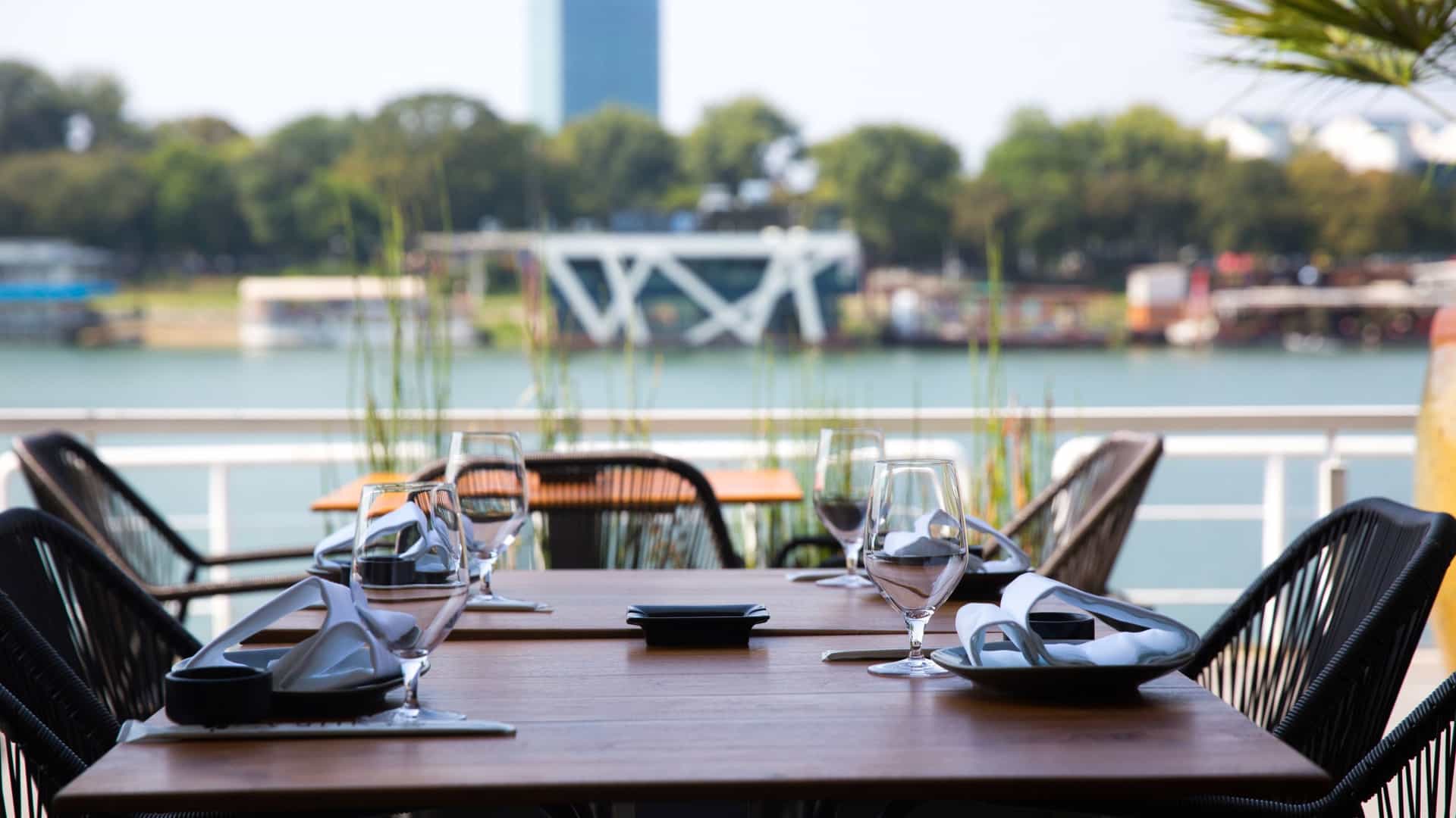 This inviting outdoor dining area, identified as a Riverside Restaurant in Saône River, features elegantly set modern wicker chairs and wooden tables. The terrace offers a stunning view of the river, a modern bridge, and a contemporary high rise building against a backdrop of lush greenery.
