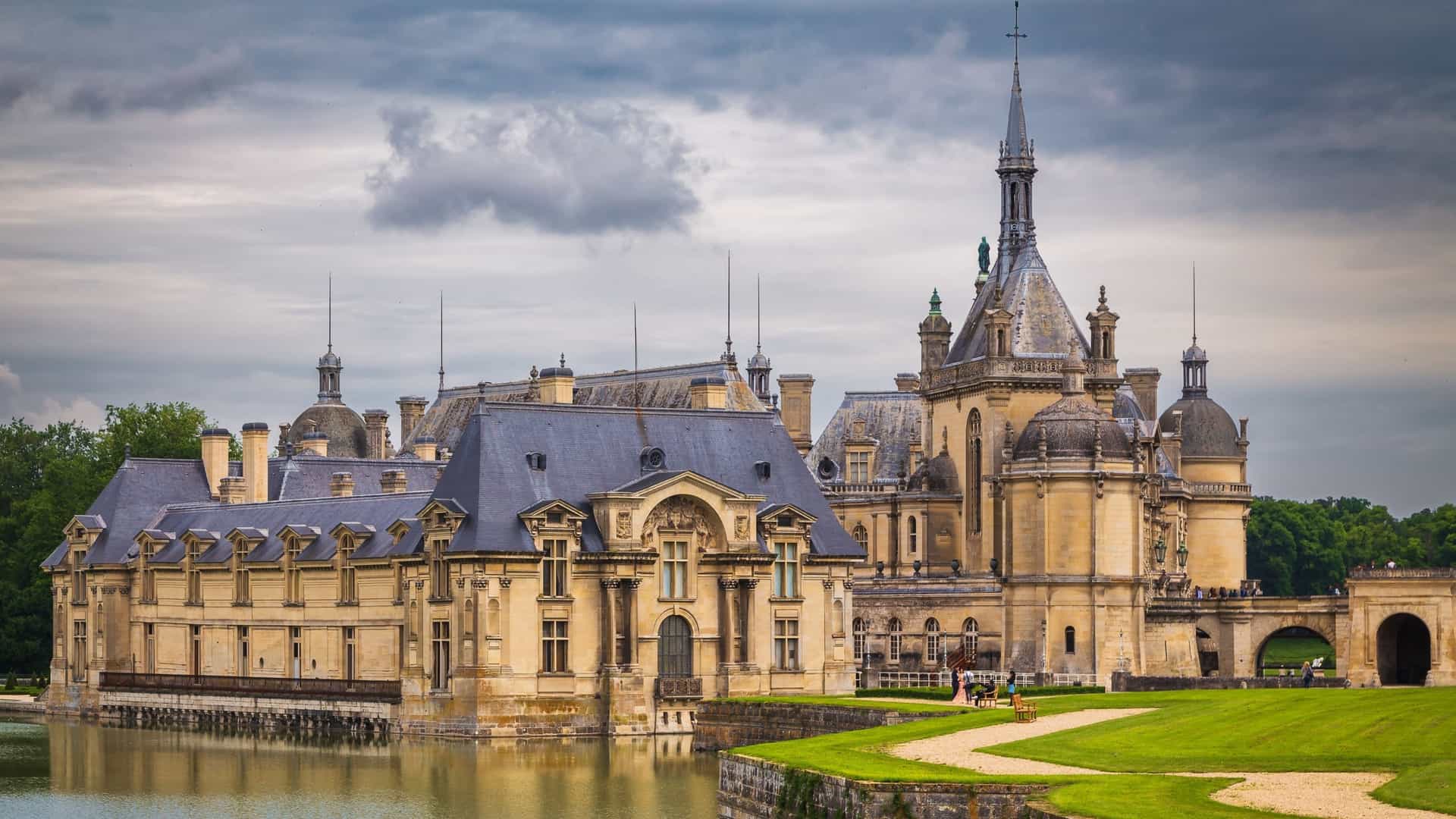 The Domaine de Chantilly, a historic French château located in the town of Chantilly, is reflected in the surrounding water. The image shows the iconic architecture of the Grand Château, which houses the Musée Condé, and the Petit Château, set amidst the vast gardens and waterways designed by André Le Nôtre.