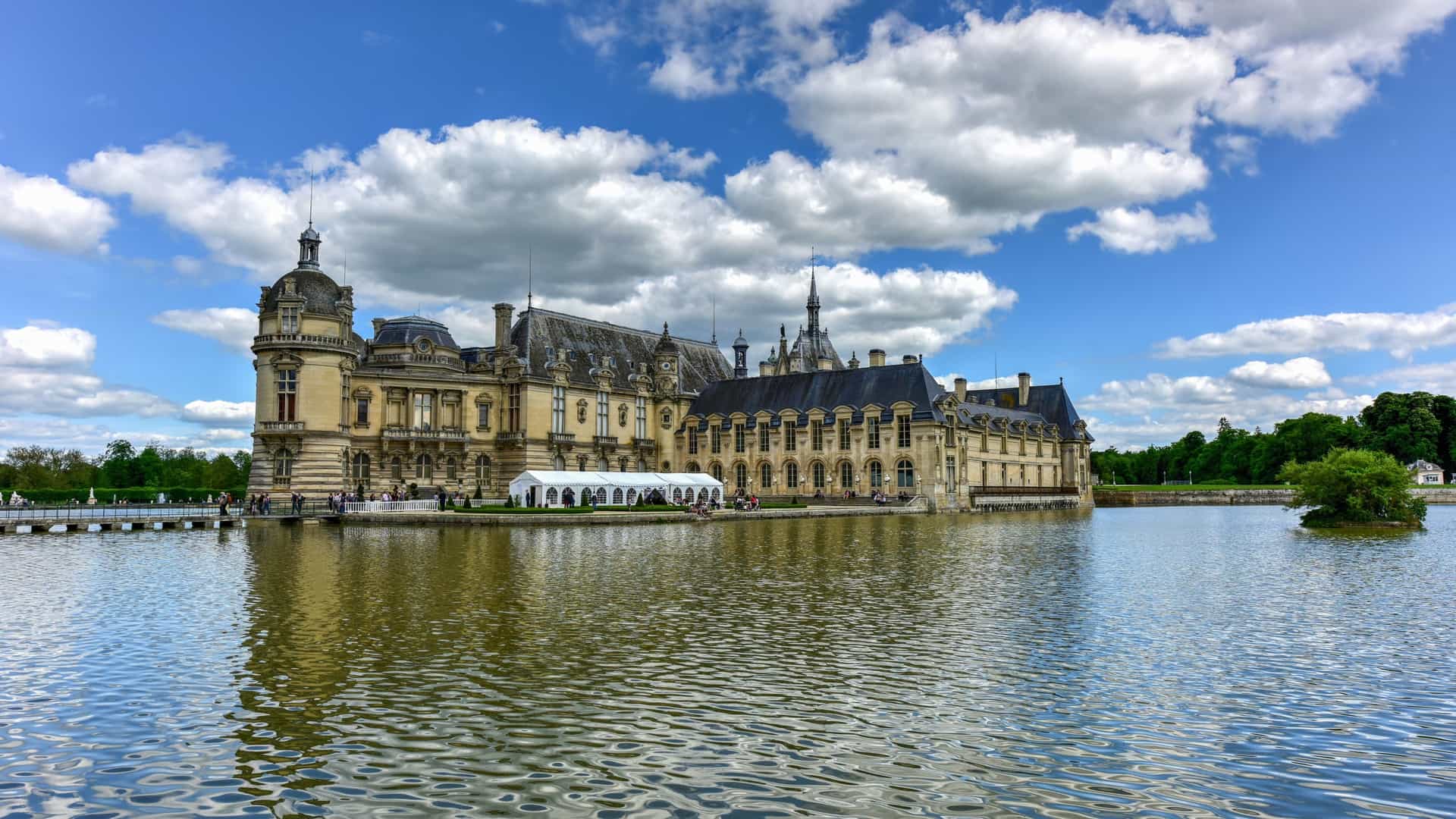A view of the Château de Chantilly, a historic French castle with towers and a moat, surrounded by a large landscaped garden.