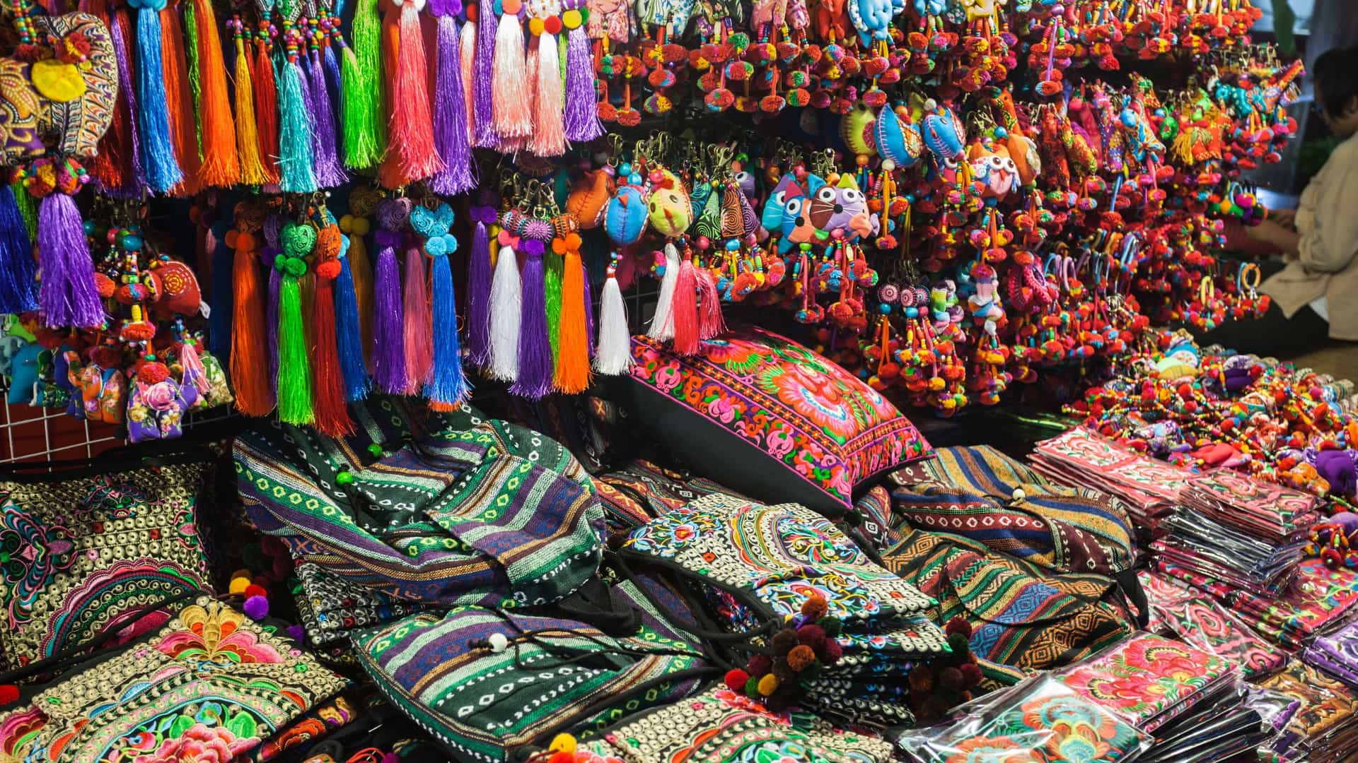 A view of a colorful Thai fabric market, with bolts and rolls of various textiles in vibrant colors and intricate patterns arranged for display and sale.