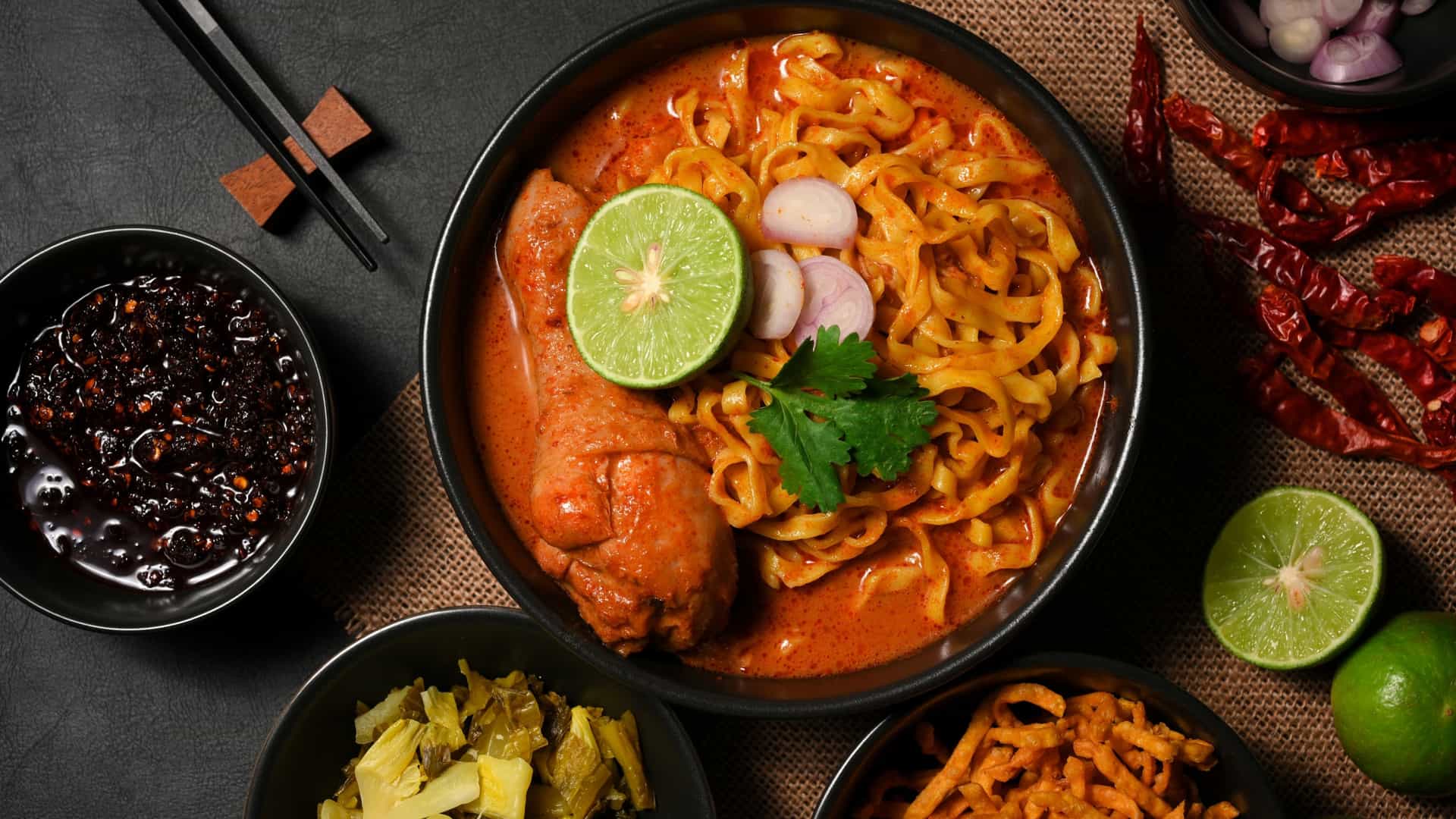 A bowl of Khao Soi, a Northern Thai coconut curry noodle soup. The rich, yellow curry broth contains tender chicken and soft egg noodles, and is topped with a nest of crispy fried noodles. The dish is garnished with fresh ingredients like sliced shallots and a lime wedge.