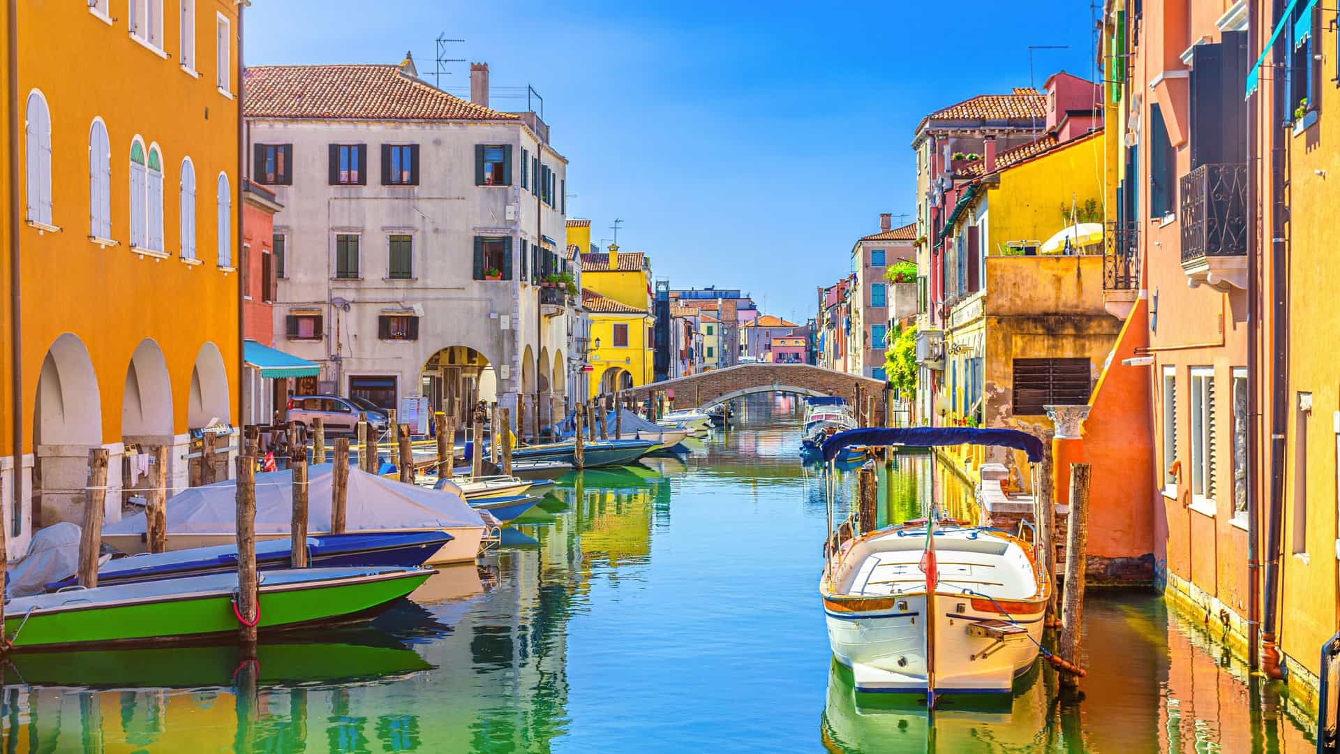 A vibrant cityscape view of Chioggia, Italy, showcasing the narrow Canal Vena lined with traditional buildings and spanned by a historic brick bridge. Multicolored fishing and leisure boats are moored along the canal, reflecting the city's maritime heritage under a clear sky.