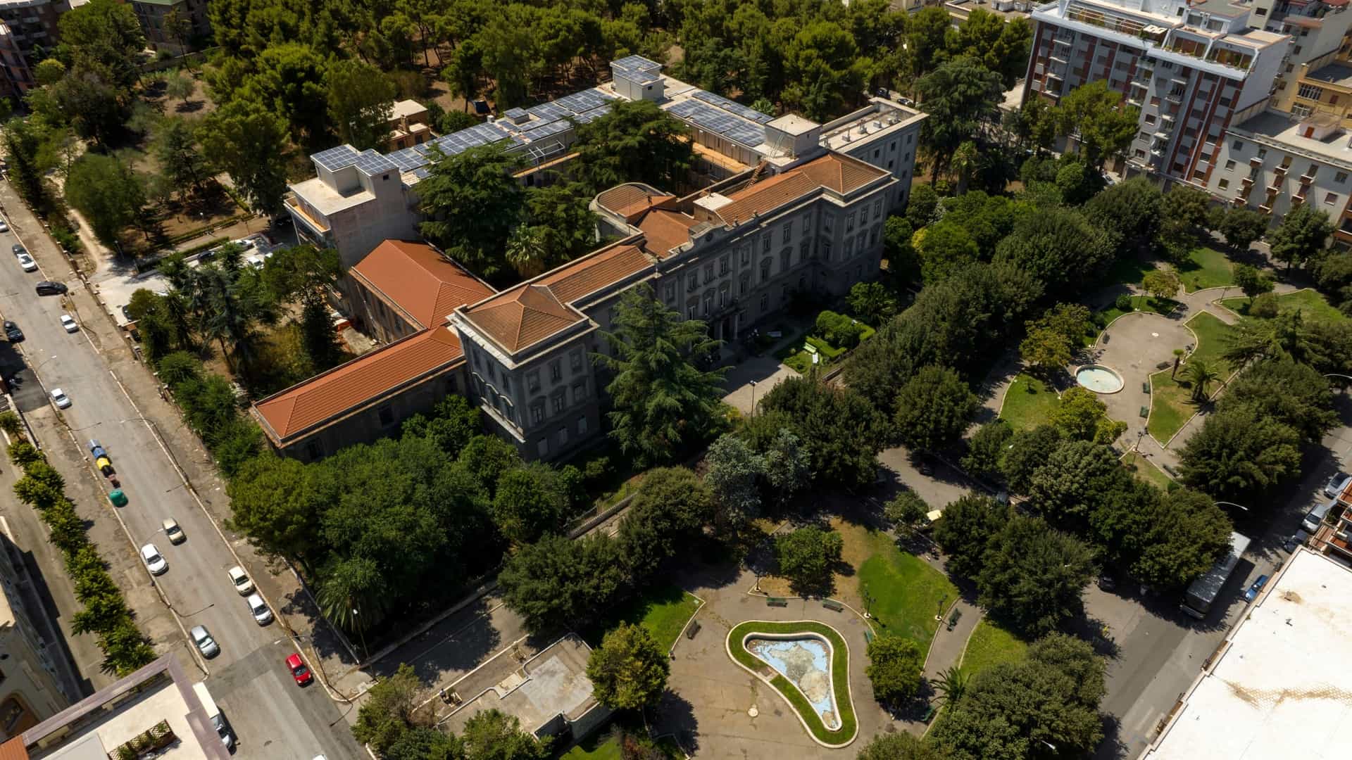 An aerial view of a large public park in Foggia, Puglia, Italy, featuring meticulously patterned green lawns, walking paths, and densely planted trees, creating a geometric and well-manicured urban green space.