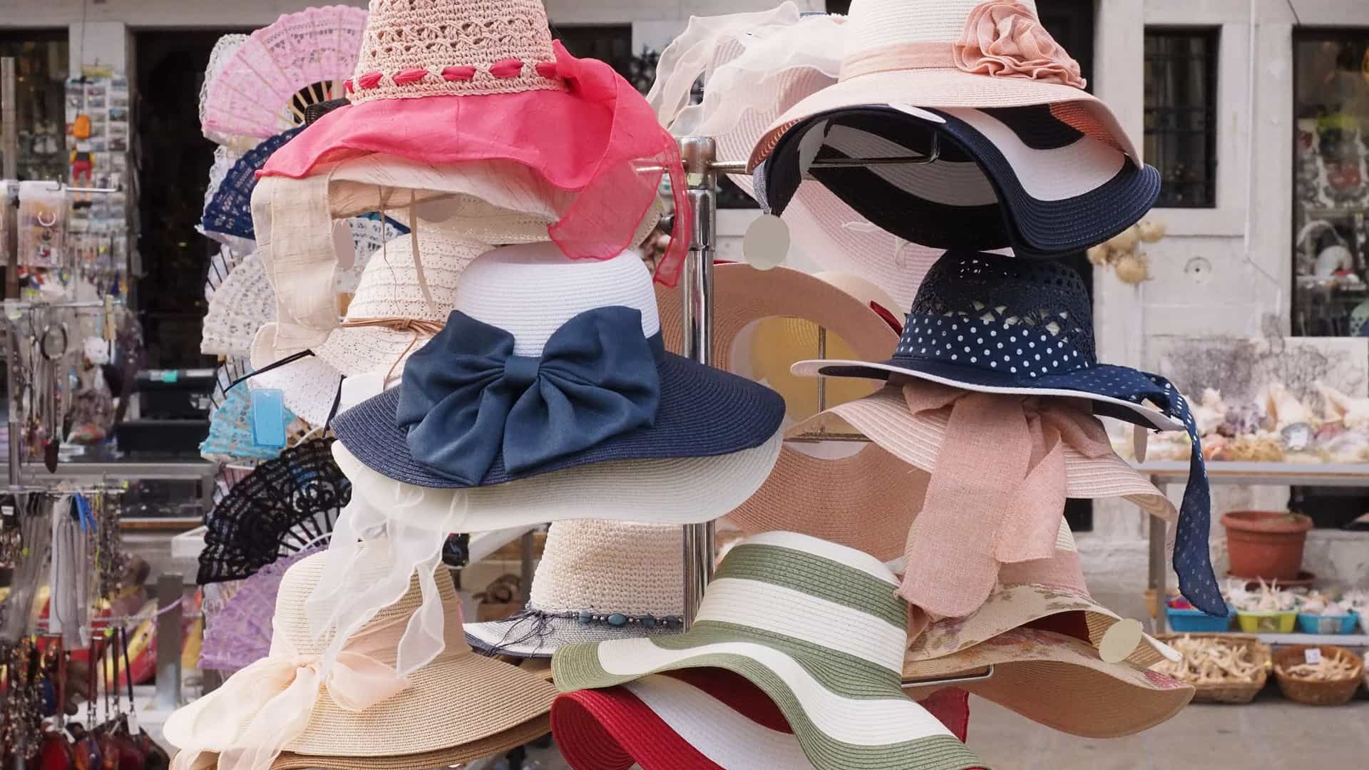 A close up shot of a street market stall displaying rows of brightly colored, woven hats for sale in Chioggia, Italy. The hats feature wide brims and various patterns in shades of red, blue, pink, yellow, and green, hanging against a dark backdrop.