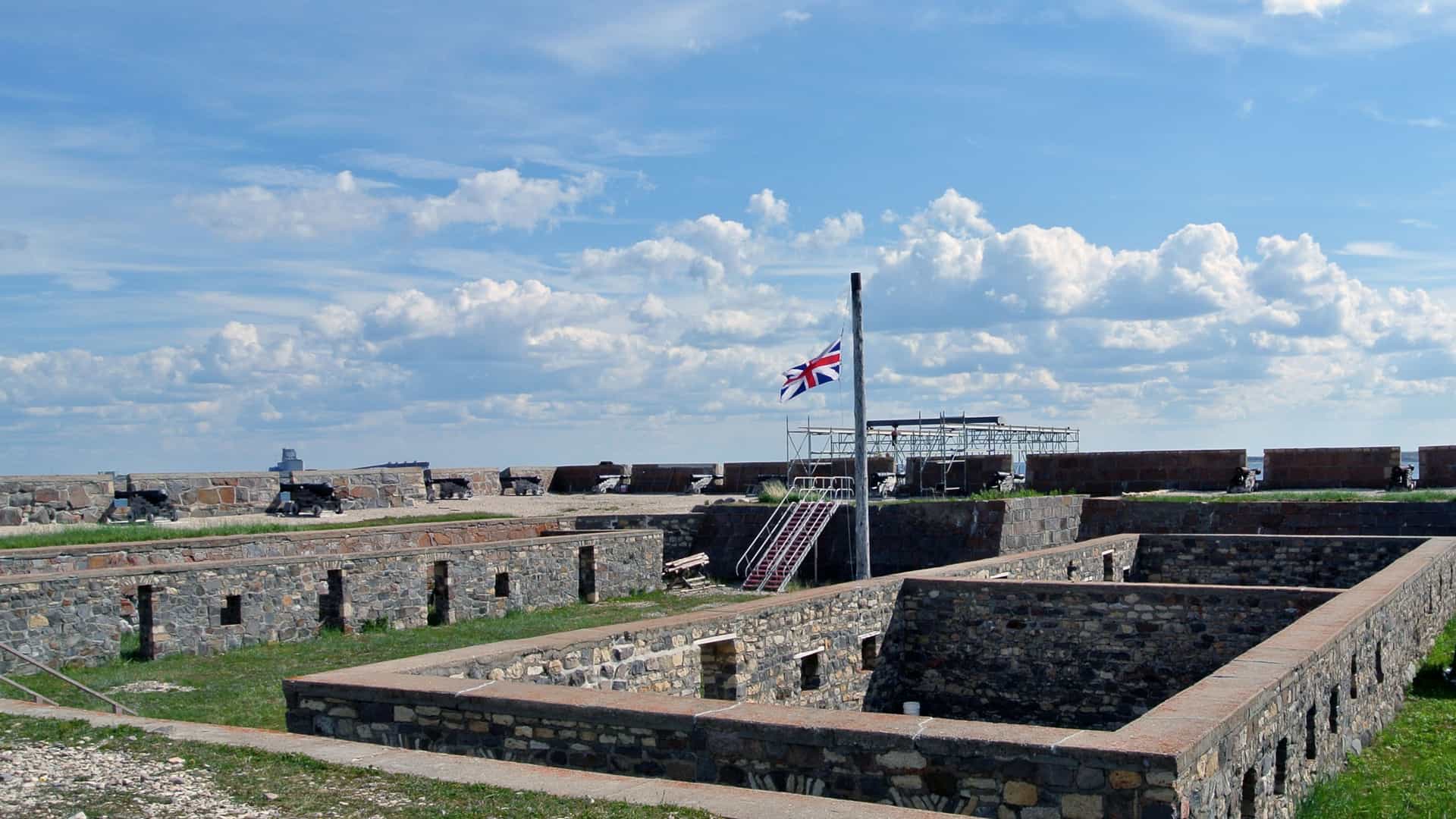 A view of the interior courtyard of the 18th century Prince of Wales Fort in Churchill, Manitoba, showcasing the imposing height and thickness of the fort's massive stone walls. The foreground features an old iron cannon resting on a stone carriage, with the bleak, flat, snow dusted tundra visible over the weathered stone battlements under a gray sky.