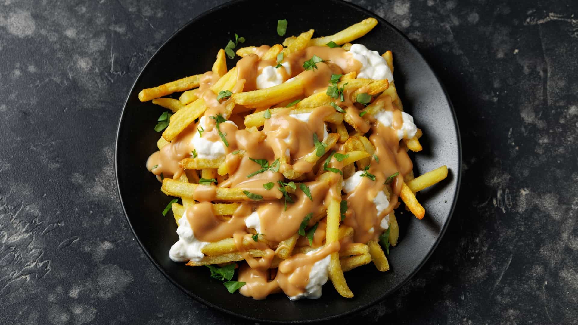 A close up view of a serving of traditional poutine, featuring a pile of golden brown french fries topped with generous chunks of squeaky white cheddar cheese curds. The dish is heavily coated in a thick, rich brown gravy that is beginning to melt the curds and is served in a black bowl.