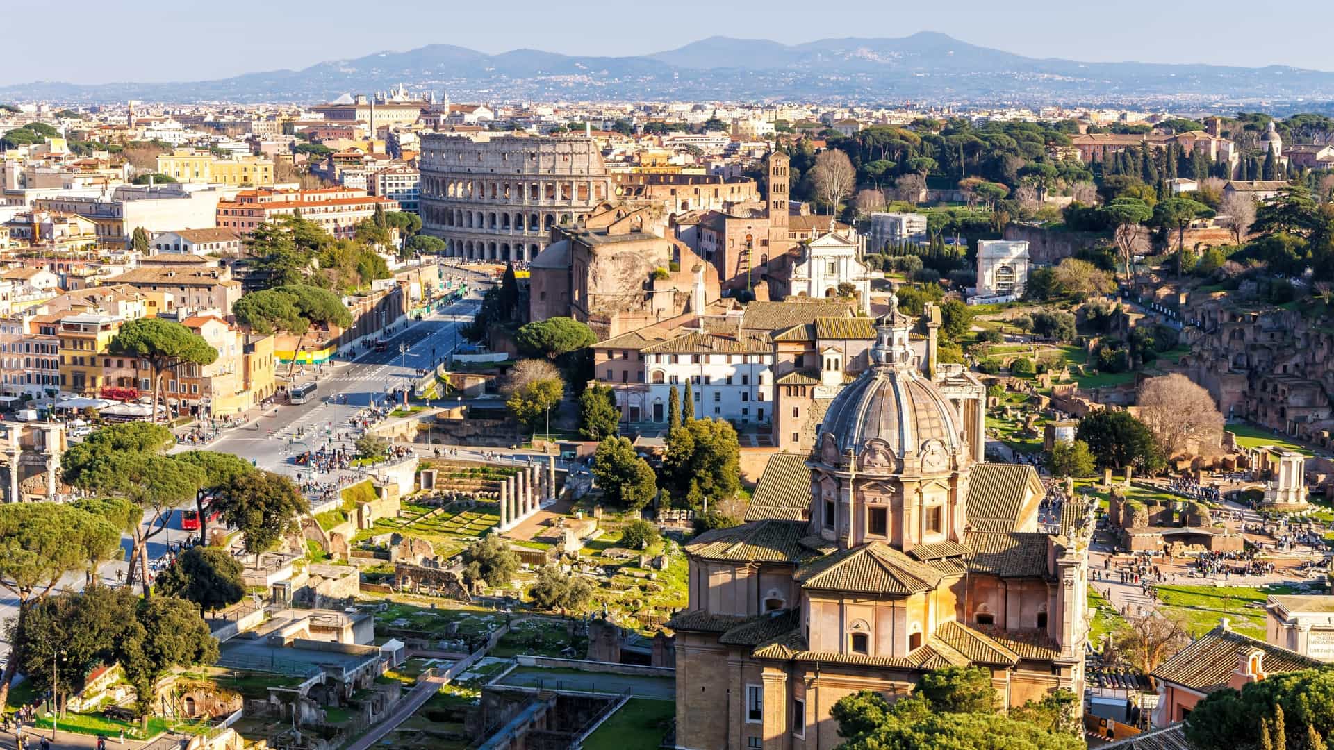 An elevated view of the historic skyline of Rome, Italy, showcasing the ancient ruins of the Roman Forum in the foreground and the iconic, elliptical structure of the Colosseum in the background. The scene is bathed in light, highlighting the historical landmarks surrounded by the modern city.