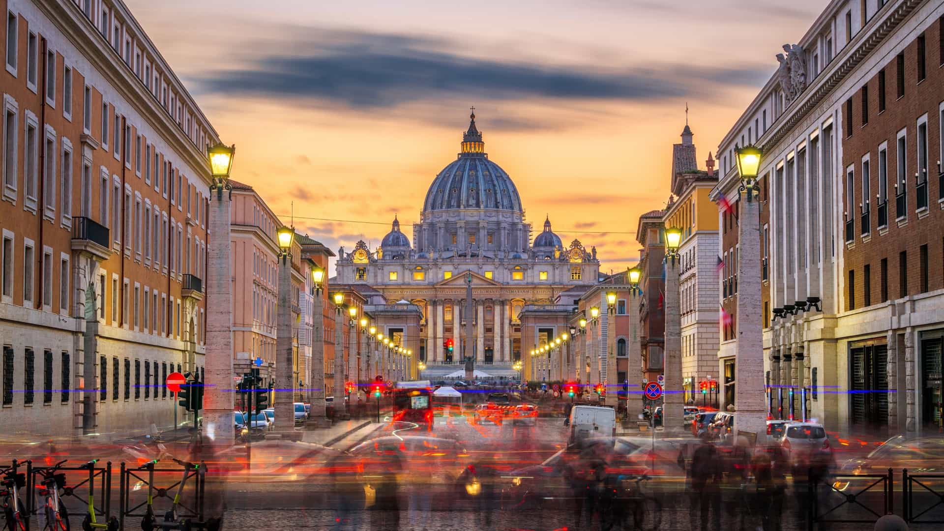 A view of Vatican City at dusk, showing the Via della Conciliazione (Road of the Conciliation) with traffic and pedestrians. The road leads towards the illuminated St. Peter's Basilica, with streetlights and buildings lining the path.