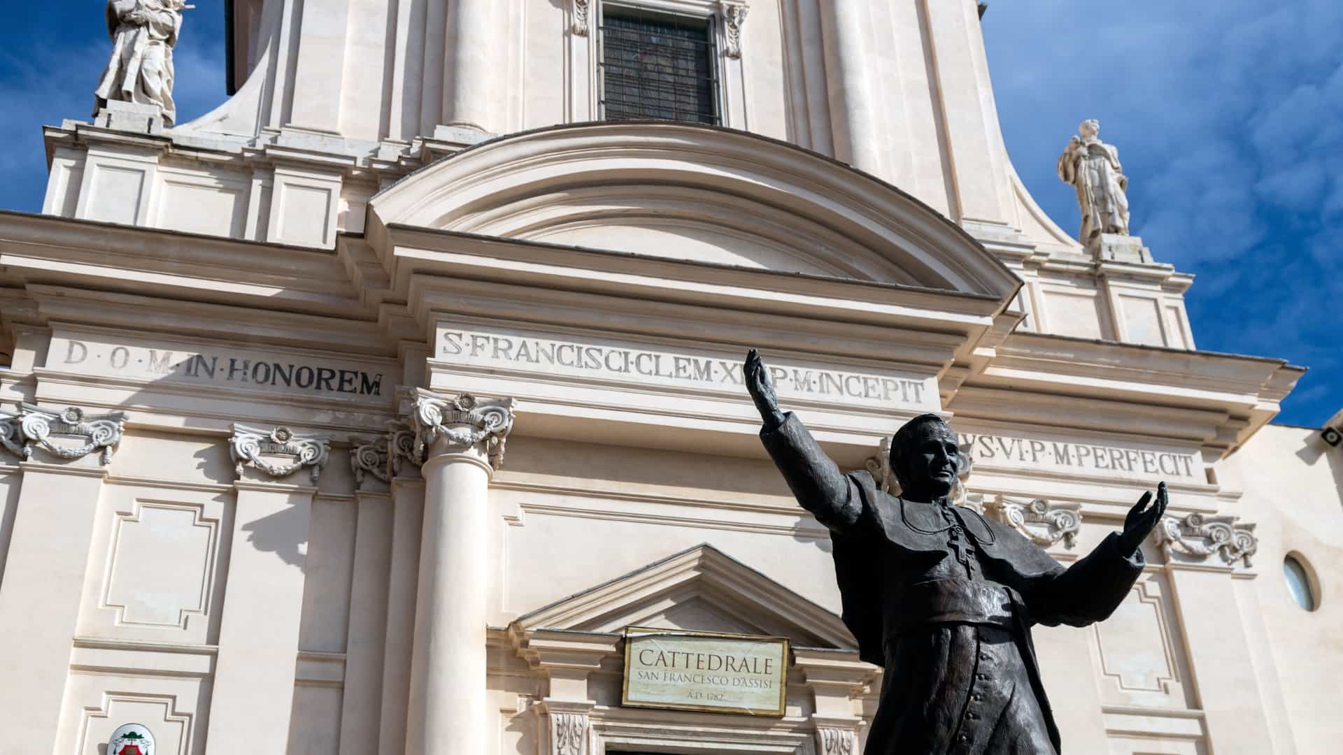 The facade of the Cathedral of San Francesco d'Assisi in Civitavecchia, Italy, a building that blends Baroque and Neoclassical architectural styles. The front of the church features two prominent statues, one of Saint Francis of Assisi and the other of Saint Anthony of Padua, on either side of a large window above the entrance.