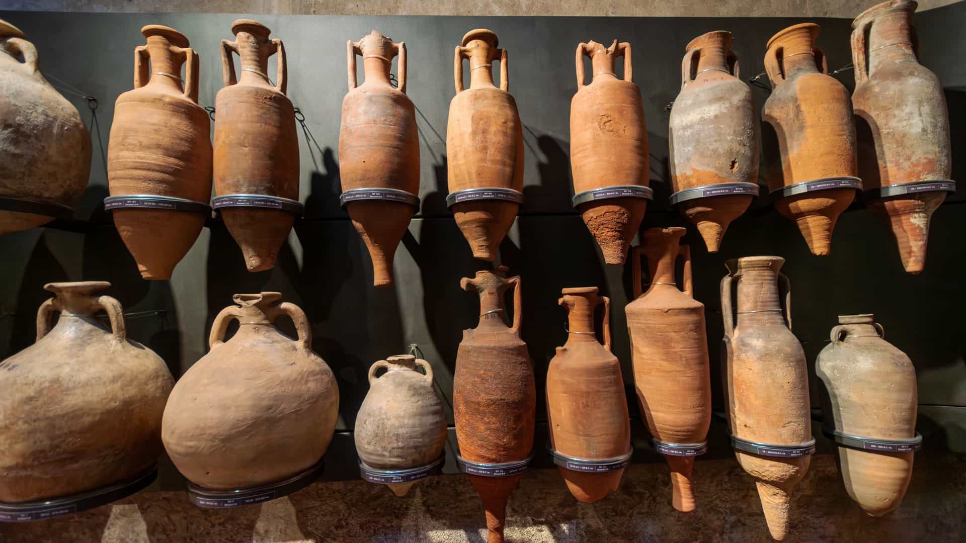 A large collection of assorted, ancient Roman amphorae (clay transport jars) on display in the National Archaeological Museum of Civitavecchia. The terracotta-colored pottery is stacked and arranged on a stone surface against a historical wall.