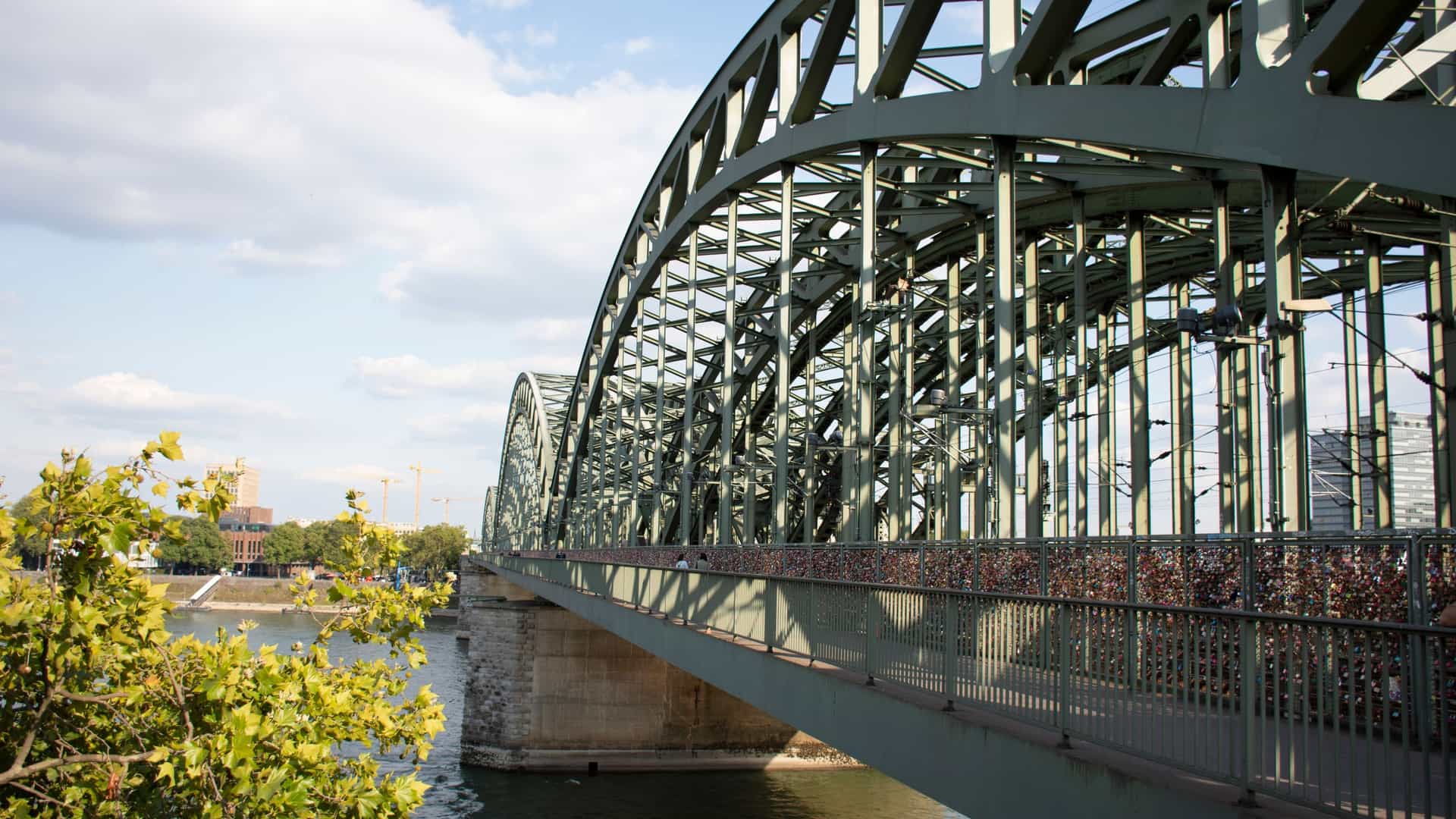 A scenic view of the Cologne cityscape in Germany, with the Rhine River flowing through the foreground. The iconic Hohenzollern Bridge, with its distinctive arch structure, spans the river, and the spires of the majestic Cologne Cathedral (Kölner Dom) and the Romanesque Great Saint Martin Church are prominent in the skyline. The river is populated with boats, and the banks are lined with a mix of historic and modern buildings.