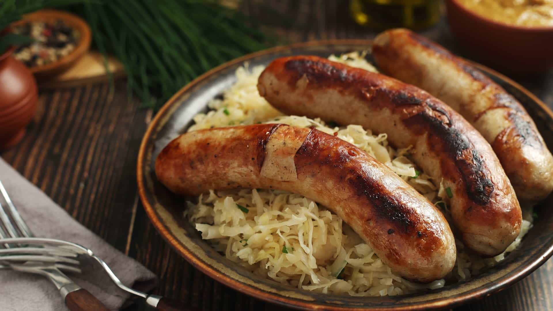 A close up of a traditional German meal, showcasing two grilled and sliced bratwurst sausages resting on a large portion of tangy sauerkraut. The savory dish is presented in a white bowl on a wooden surface, with a small side of yellow mustard.
