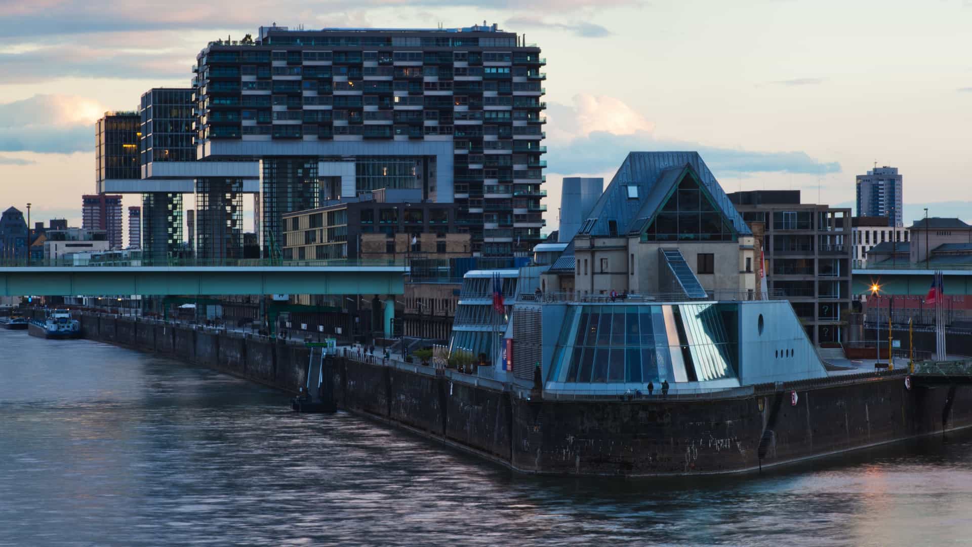 An aerial view of the Lindt Chocolate Museum in Cologne, Germany, a modern, boat shaped glass and steel structure built on a peninsula overlooking the Rhine River. In the foreground, the historic stone Malakoff Tower is visible, with the contemporary architecture of the Rheinauhafen (including the "Crane Houses") stretching along the riverbank behind it.