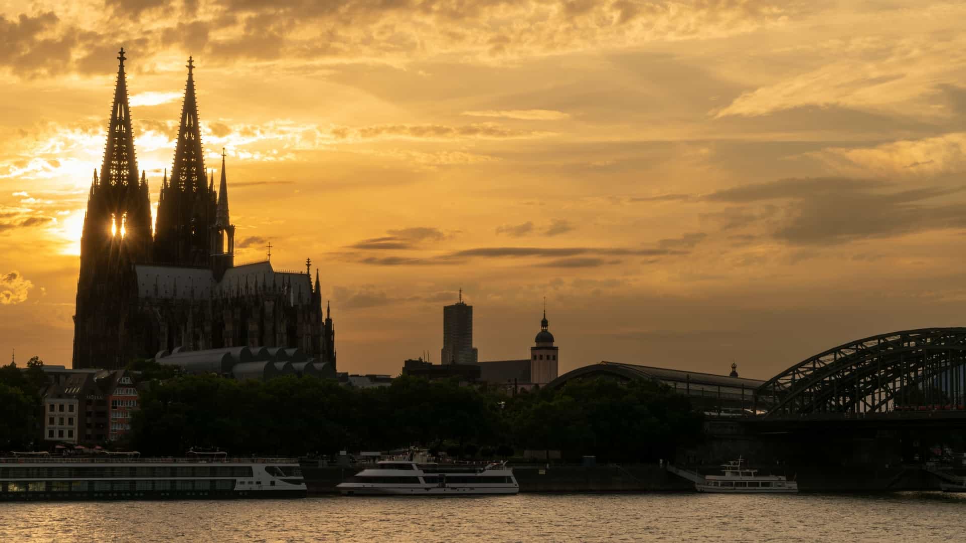 A breathtaking wide angle view of the Cologne skyline at sunset, taken from across the Rhine River. The two massive, filigreed Gothic spires of the Cologne Cathedral (Kölner Dom) dominate the center, silhouetted against a dramatic sky of fading orange, yellow, and purple hues. In the foreground, the arches of the Hohenzollern Bridge (Hohenzollernbrücke) span the river, filled with the silhouettes of passing trains and famous for its railings covered in countless "love locks." The Rhine River reflects the remaining light of the setting sun.