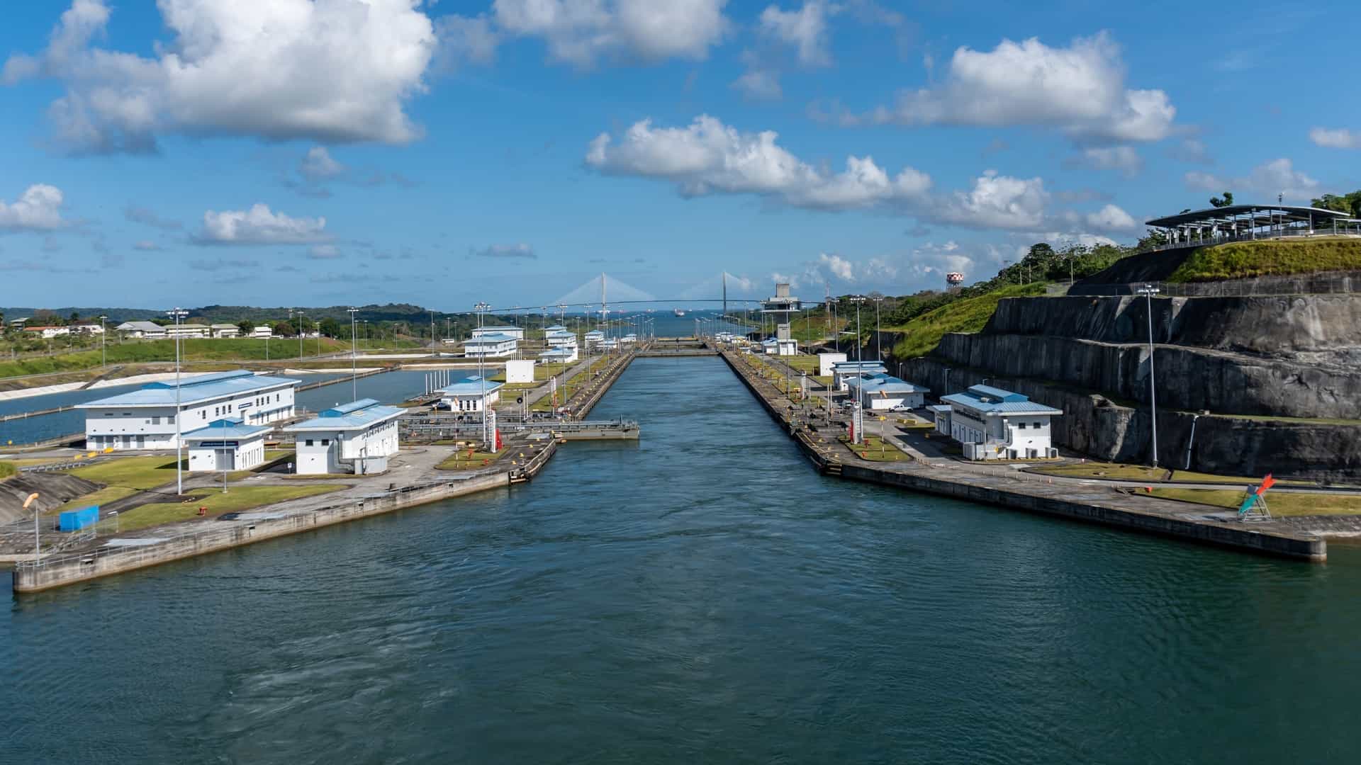 A panoramic view from the Agua Clara Locks Visitor Center in Panama shows the massive, new Neopanamax lock chambers of the Panama Canal expansion. The foreground features the modern observation deck, while a huge container ship is visible traversing the colossal concrete structure toward Gatún Lake.