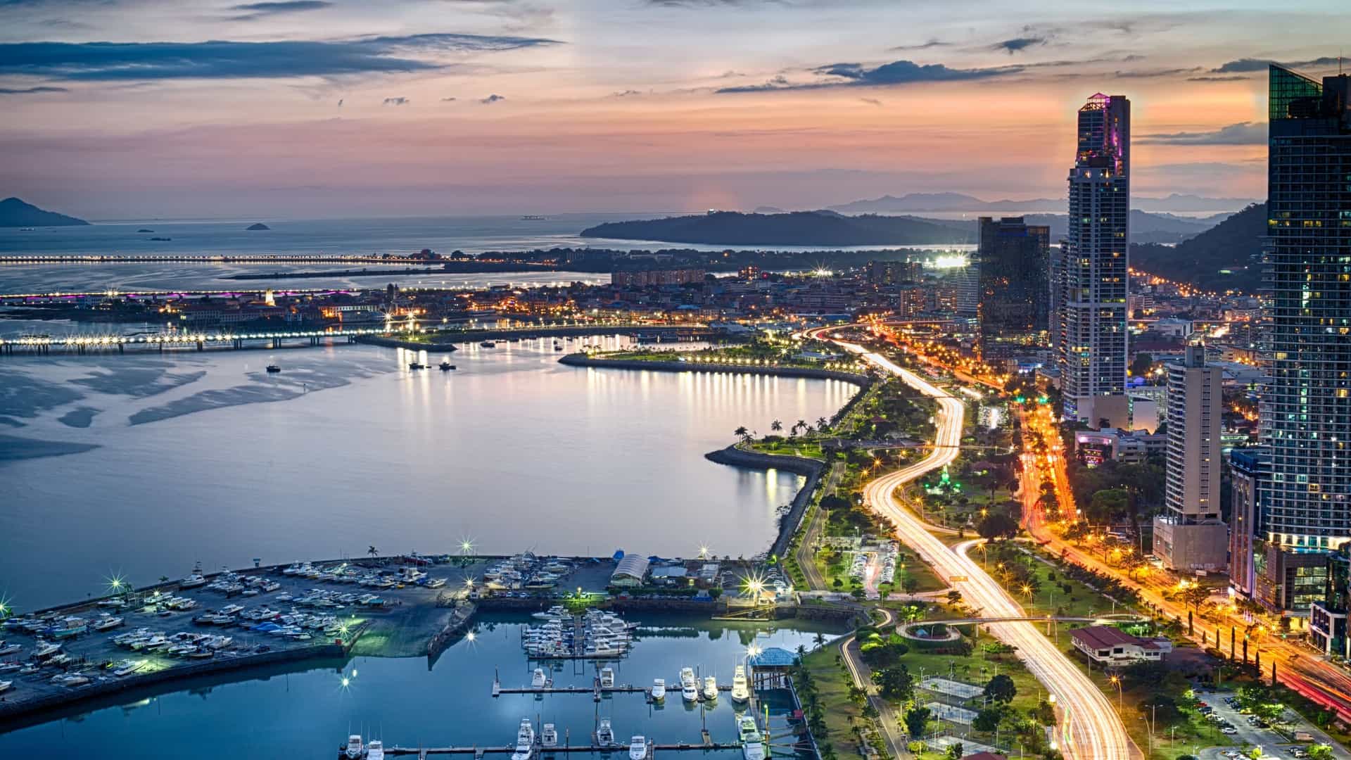 A stunning twilight view of Avenida Central in Panama City, Panama, showing the boulevard curving along the waterfront with modern skyscrapers brightly lit against the dark sky and their reflections on the bay.