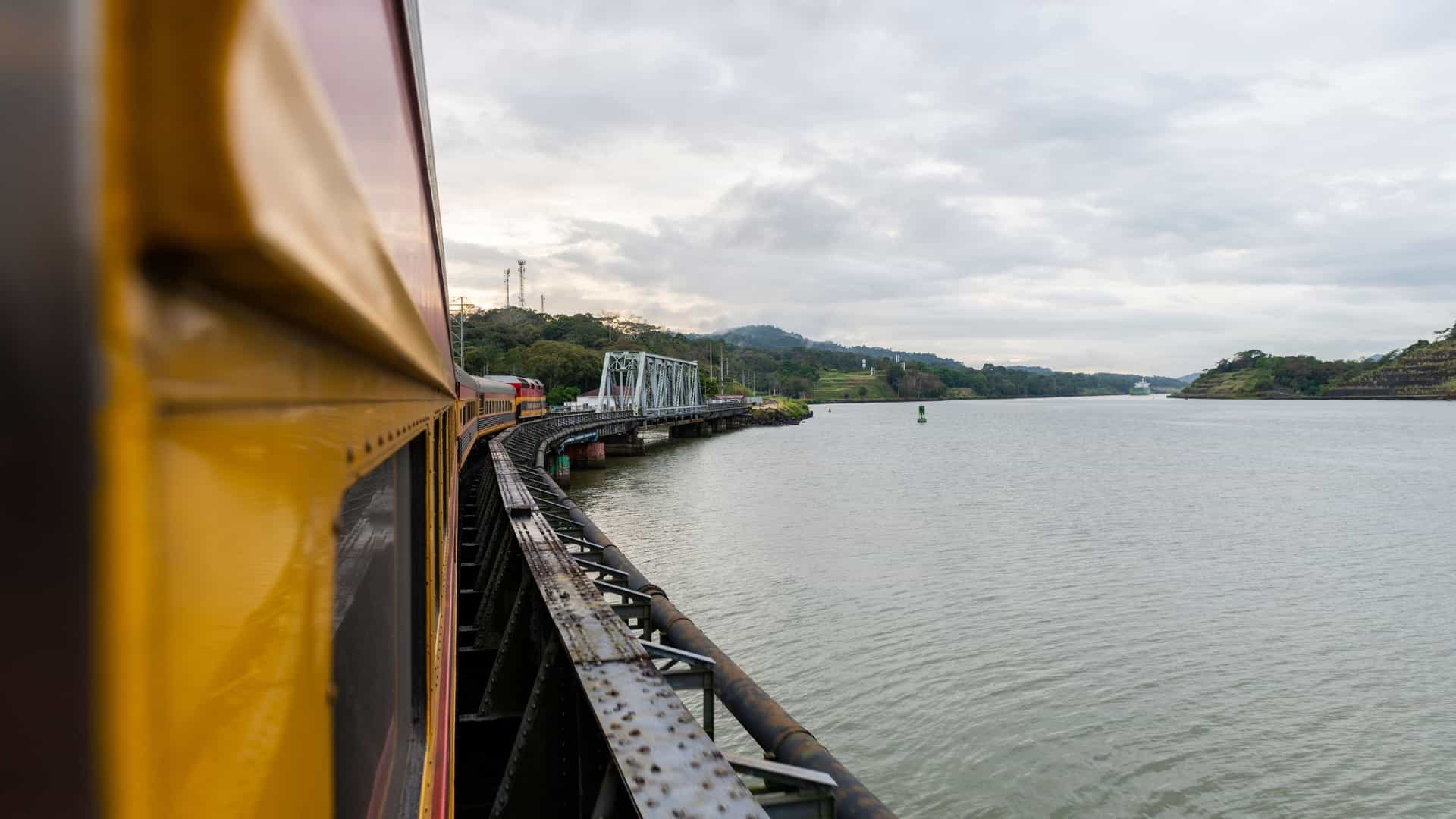 A passenger train on the Panama Canal Railway travels along the water's edge, offering panoramic views as it links the Atlantic and Pacific oceans across the Panamanian isthmus. The modern train features a distinctive gold and blue livery and is captured moving past the lush, green rainforest that borders the historic canal route.