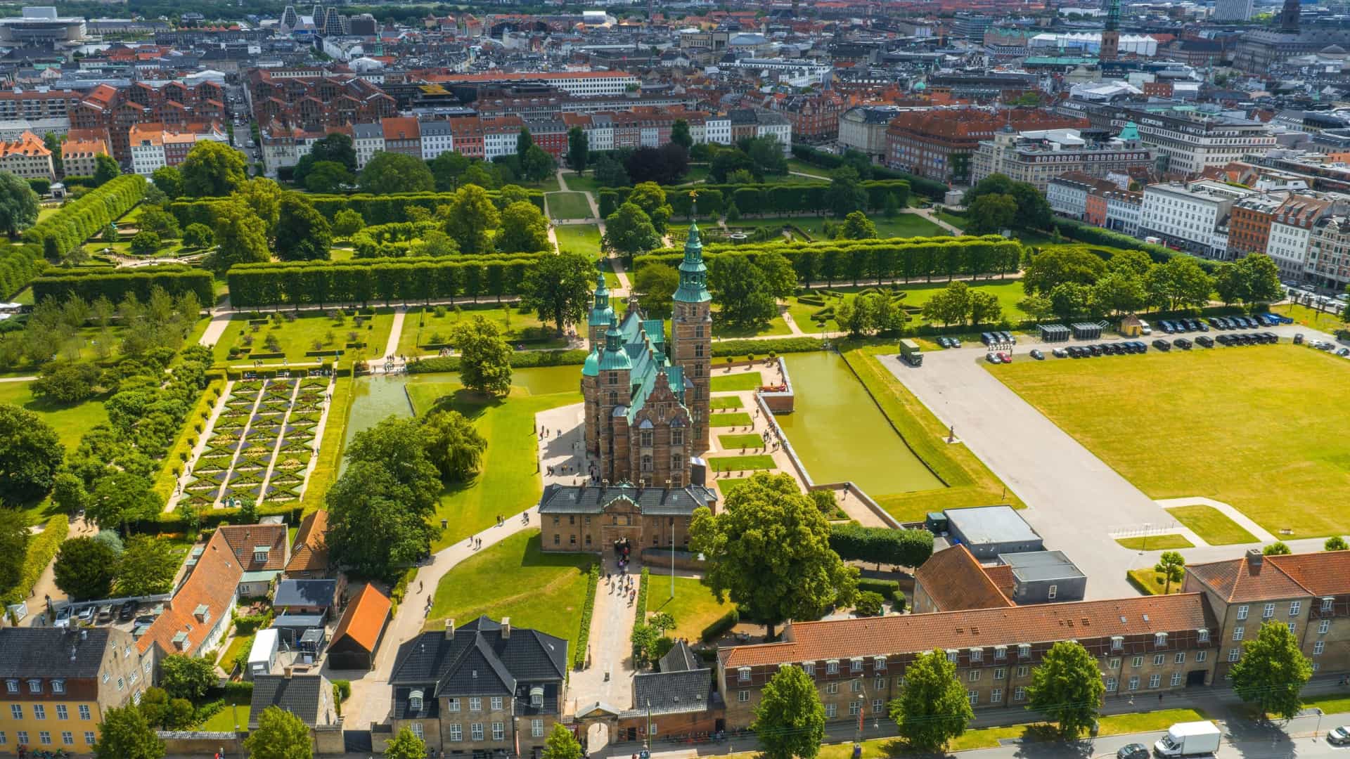 An aerial view of Rosenborg Castle in Copenhagen, Denmark, a Renaissance style building with green spires and a surrounding moat. The castle is set within the symmetrical, lush green King's Garden, with pathways visible through the trees.