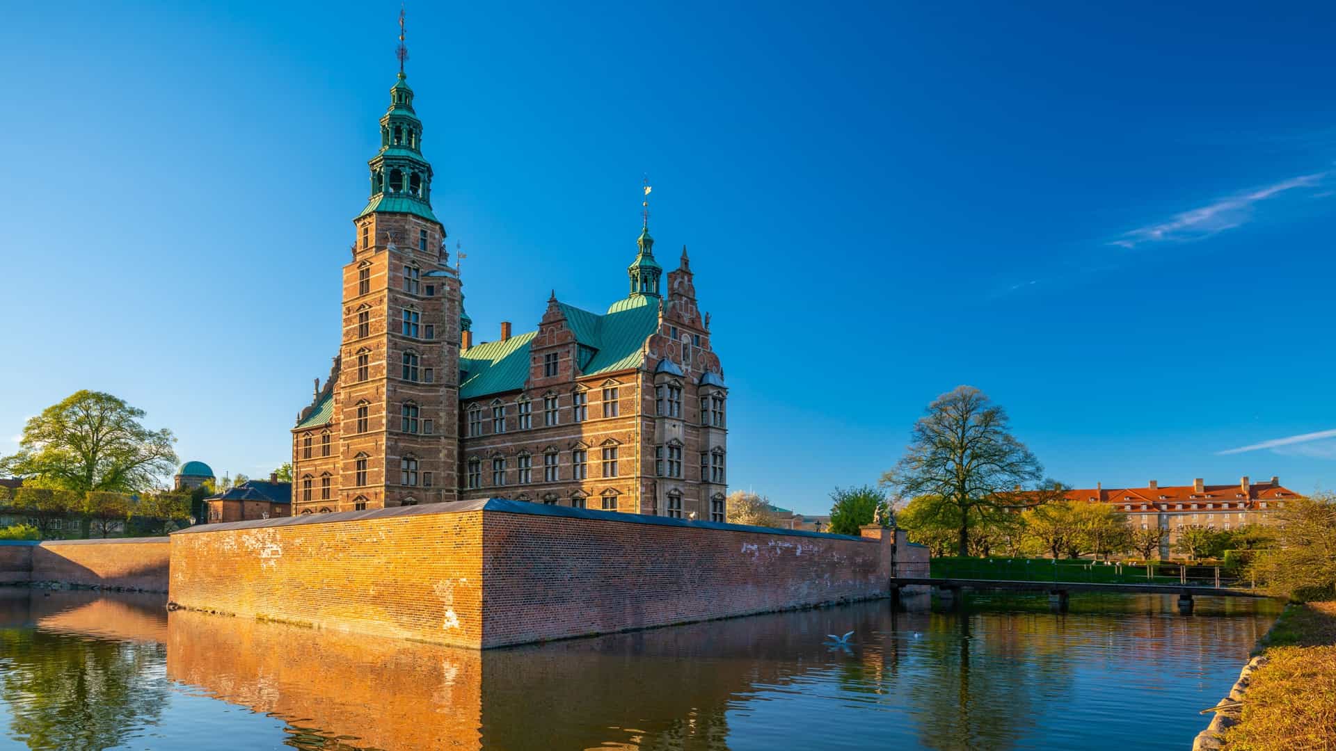 A view of Rosenborg Castle in Copenhagen, Denmark, under a clear blue sky. The Renaissance castle is visible beyond the lush green lawns and hedges of the surrounding King's Garden.