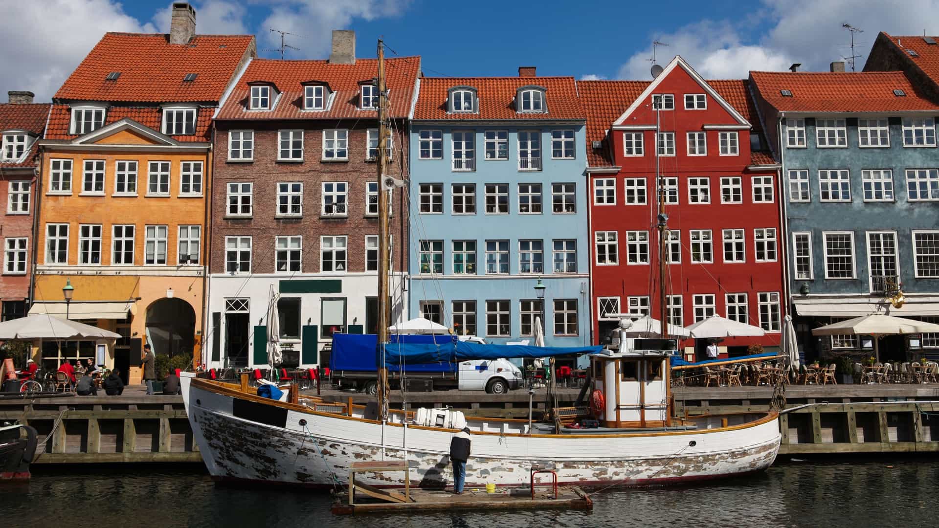 A view of Nyhavn, a historic harbor in Copenhagen, Denmark, with a row of brightly colored townhouses lining the canal. Several classic wooden ships are docked along the waterfront.