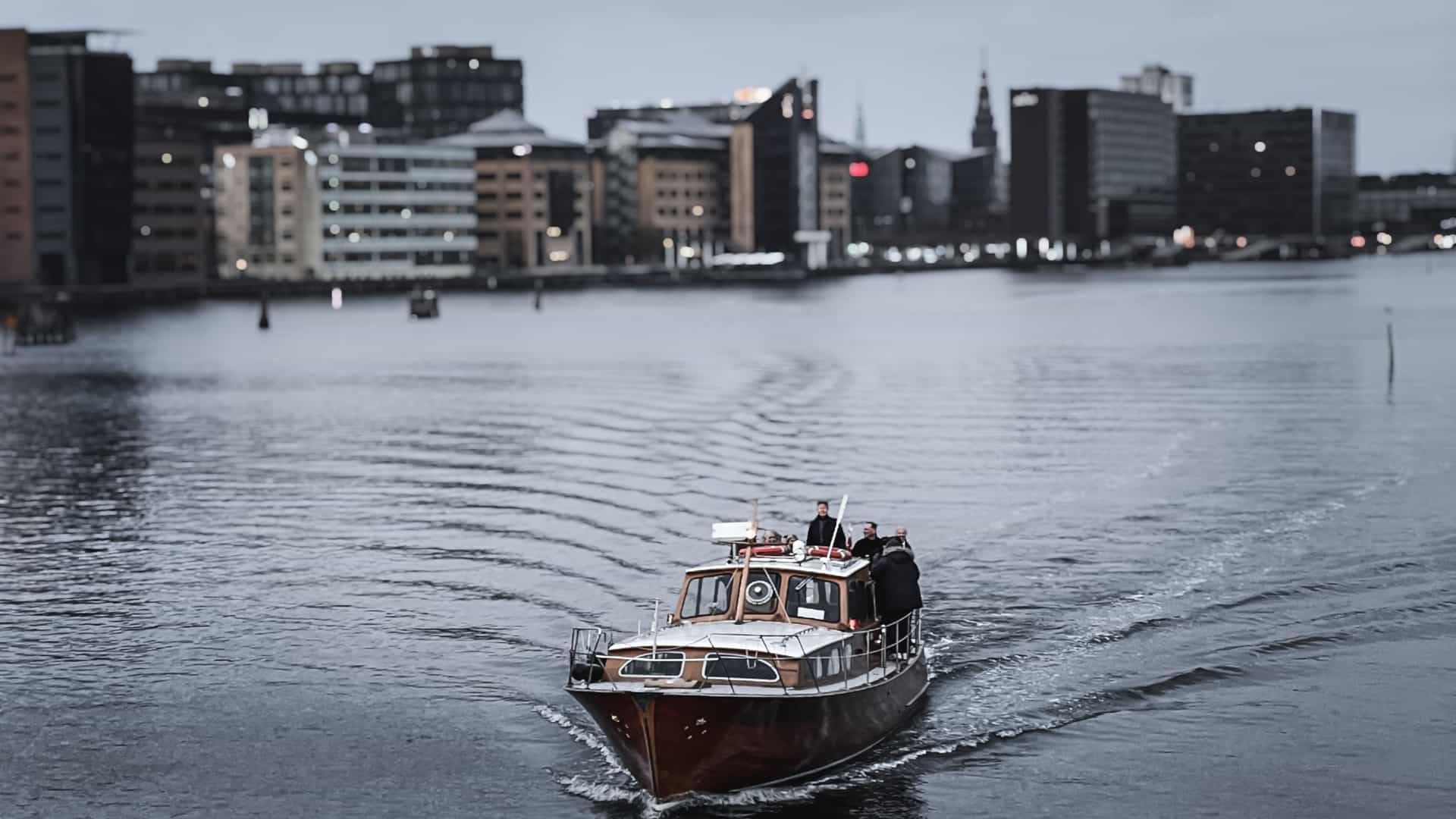 A classic canal cruise boat with tourists on board sails down a Copenhagen canal, passing a line of brightly colored, historic townhouses on the left, likely in the Nyhavn district. The water is calm, and the scene is characterized by the city's blend of historic architecture and maritime activity.