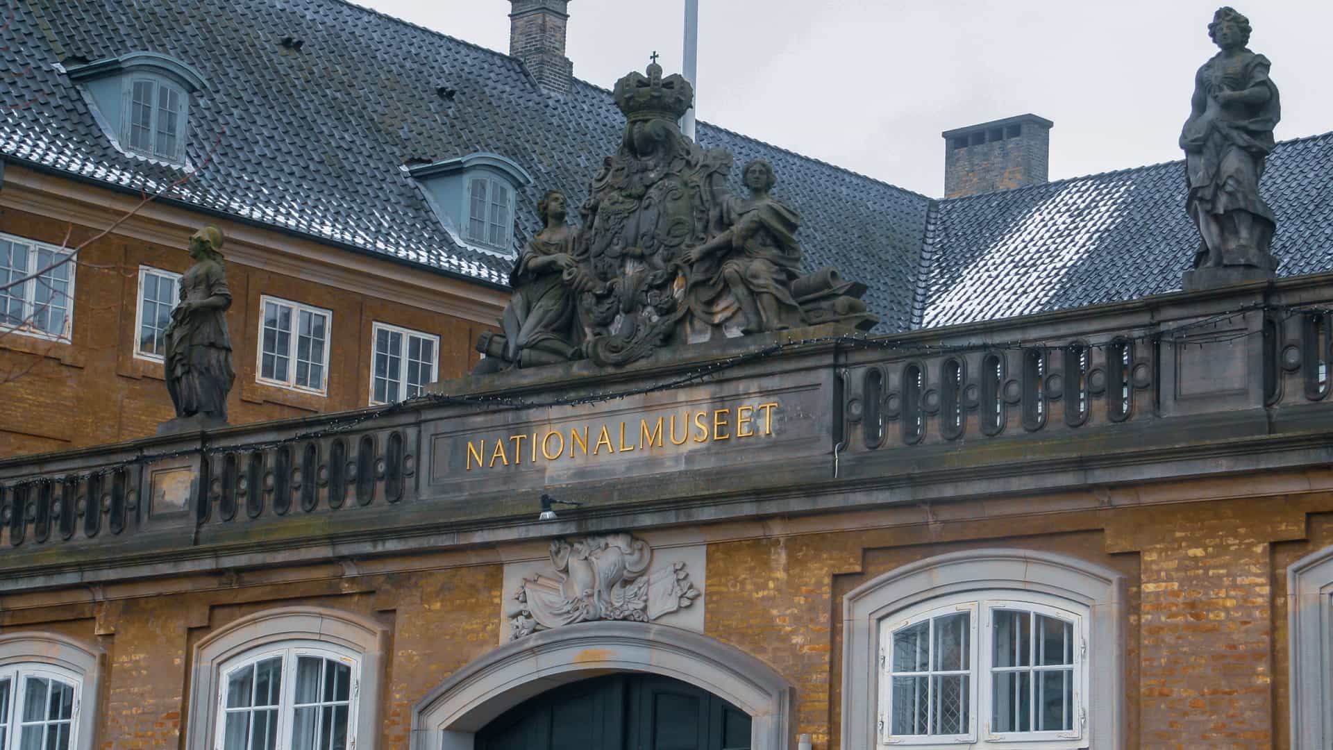 A close up view of the entrance to the National Museum (Nationalmuseet) in Copenhagen, Denmark, a historic brick building with a large golden sign over the door. The roofline is decorated with a balustrade and several large sculptures, including a central, ornate royal crest and two figures standing on either side.