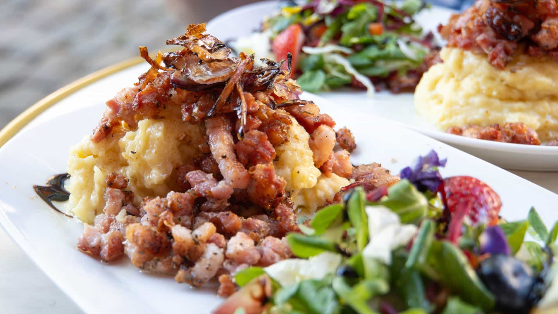 A rustic, close up view of the traditional Danish dish "Brændende Kærlighed" (Burning Love) served on a white plate. It consists of a large mound of creamy, buttered mashed potatoes with a well in the center, which is filled with a mixture of crispy fried bacon pieces and soft, caramelized onions. The dish is typically garnished with fresh herbs, such as chopped parsley or chives, and often accompanied by slices of pickled beets.