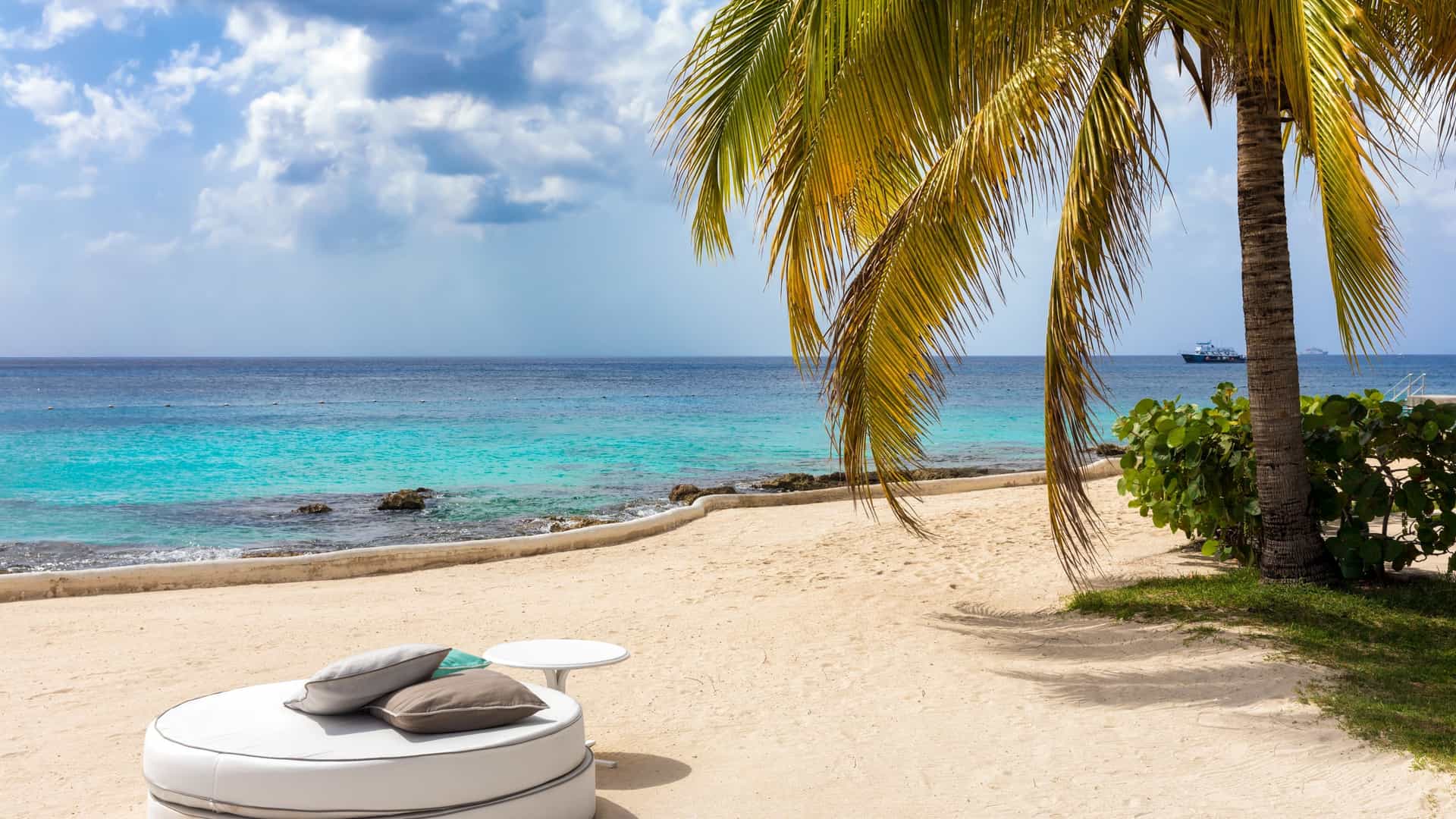 A luxurious sunbed with pillows rests on a white sand beach under the shade of palm trees. In the background, the turquoise water of the Caribbean Sea is visible. The scene is located on Cozumel Island, Mexico.