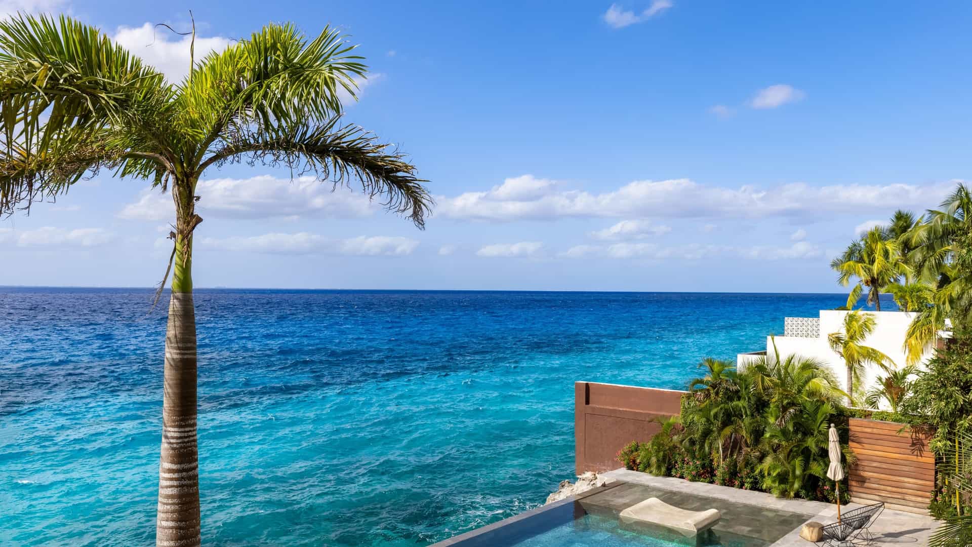 A sunny scene capturing the waterfront of San Miguel de Cozumel, Mexico, with the vibrant Caribbean Sea and a clear blue sky dominating the background. The foreground likely features the coastal promenade (Avenida Rafael E. Melgar), which is lined with colorful buildings and shows the nearby presence of ferry docks or cruise ship piers.