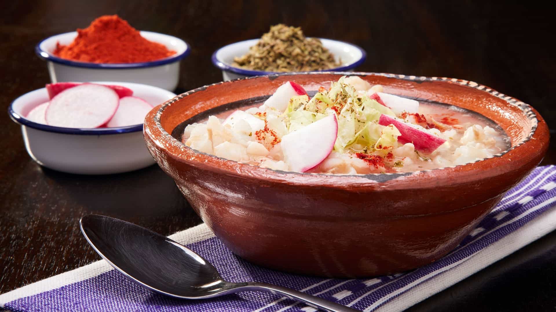 A vibrant, overhead view of a steaming bowl of Pozole Blanco (White Pozole), a traditional Mexican soup. The rich, clear white broth contains large, plump kernels of hominy and generous shreds of tender chicken. The soup is heavily garnished with crisp, freshly sliced toppings, likely including shredded cabbage, thin radish slices, chopped onion, and a lime wedge on the side.