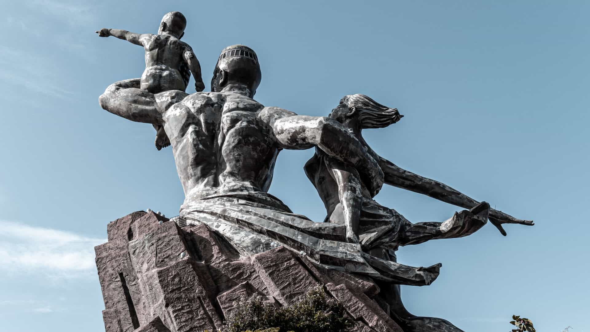 A ground level shot of the African Renaissance Monument in Dakar, Senegal, emphasizing the backside of the colossal bronze statue. The view focuses on the muscular back of the man and the flowing drapery and hair of the woman as they emerge from the base, with the sky or hillside serving as the background.