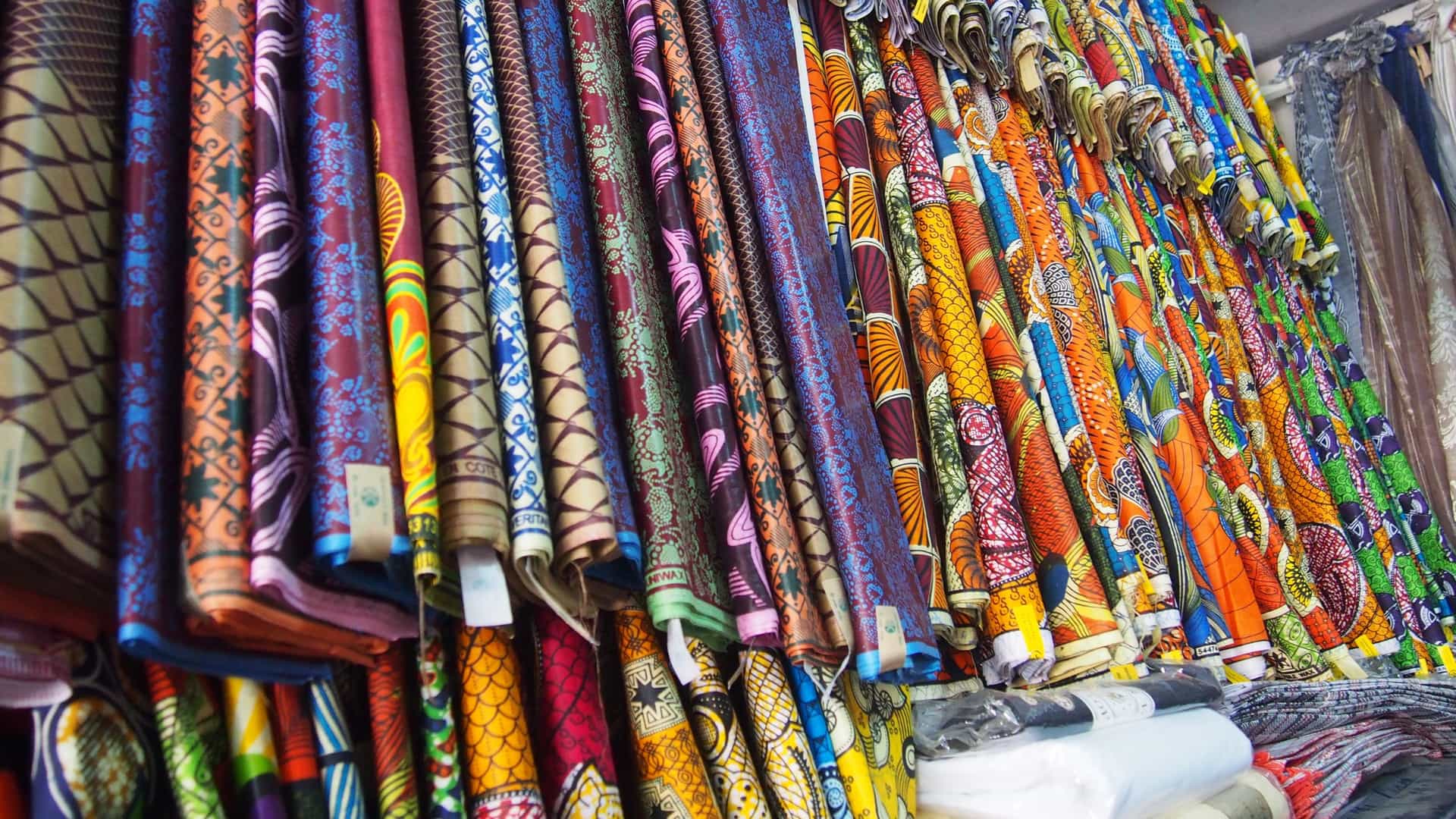 A vibrant close up of a textile stall, showcasing dozens of rolled bolts of African wax print fabric (Ankara) stacked floor to ceiling, likely photographed in Marché Sandaga in Dakar, Senegal. The display features a dense array of complex geometric and floral patterns in a rich spectrum of colors, including blues, reds, yellows, and purples.
