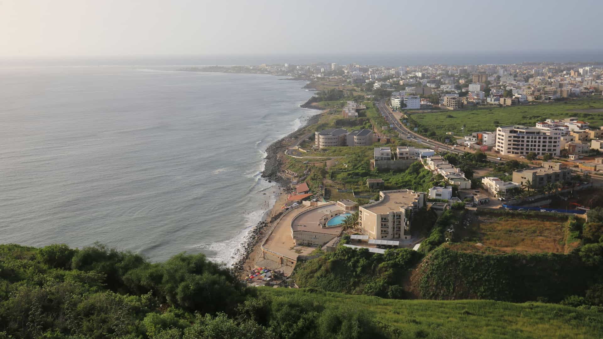 A high angle panoramic view taken from the Les Mamelles Lighthouse (Phare des Mamelles) on the Cap Vert peninsula in Dakar, Senegal. The image captures the Atlantic Ocean coastline and the suburban area known as Pointe des Almadies. In the distance, the cityscape of Dakar features modern buildings, beaches, and the rocky shoreline, all under a bright sky. The view includes the expansive ocean meeting the westernmost point of the African continent.