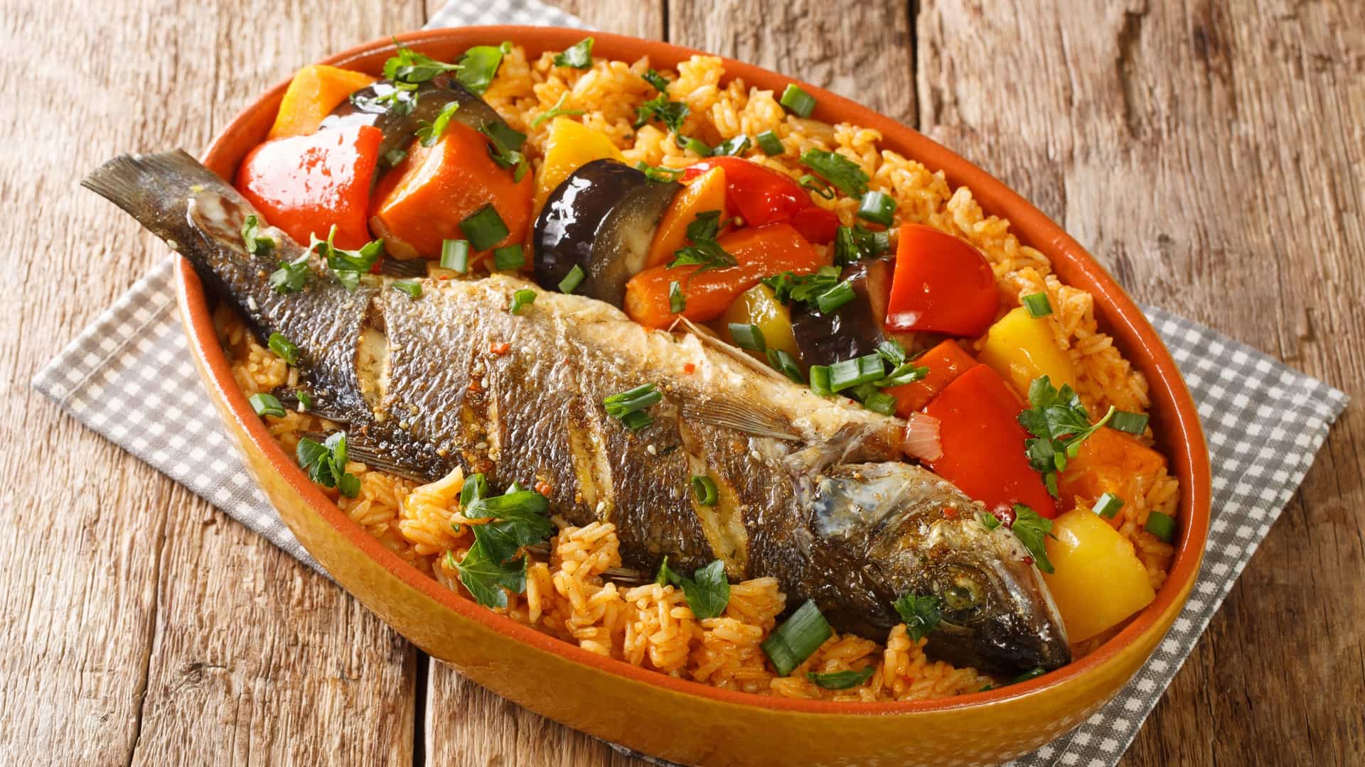 A close up, overhead shot of a platter of Thieboudienne, the national dish of Senegal. The plate is filled with rice cooked in a red tomato sauce, and topped with pieces of fish and an assortment of vegetables, including carrots and cabbage.
